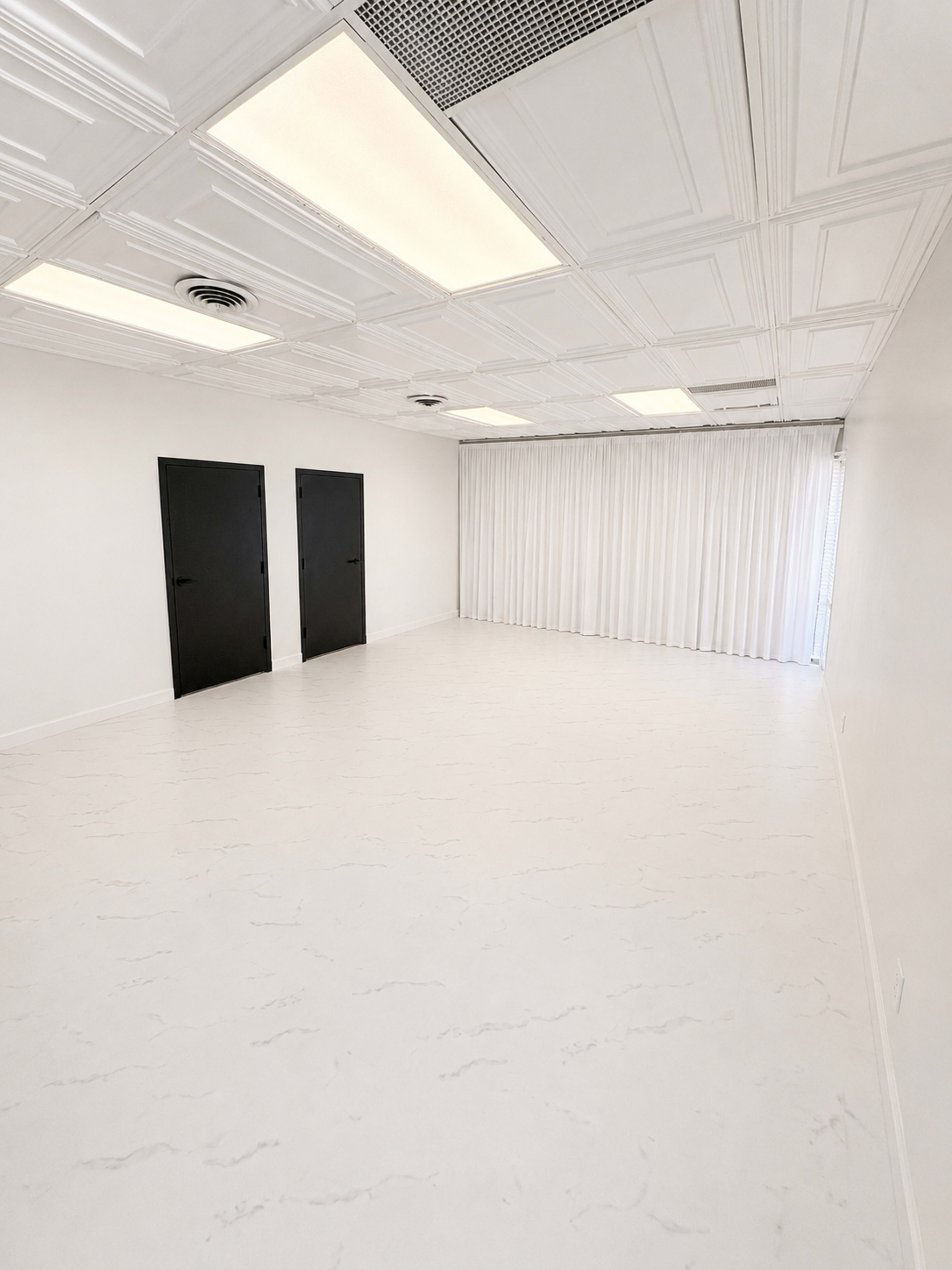 The image shows a spacious, empty room with white walls and flooring, featuring two black doors and a row of curtains on one side.