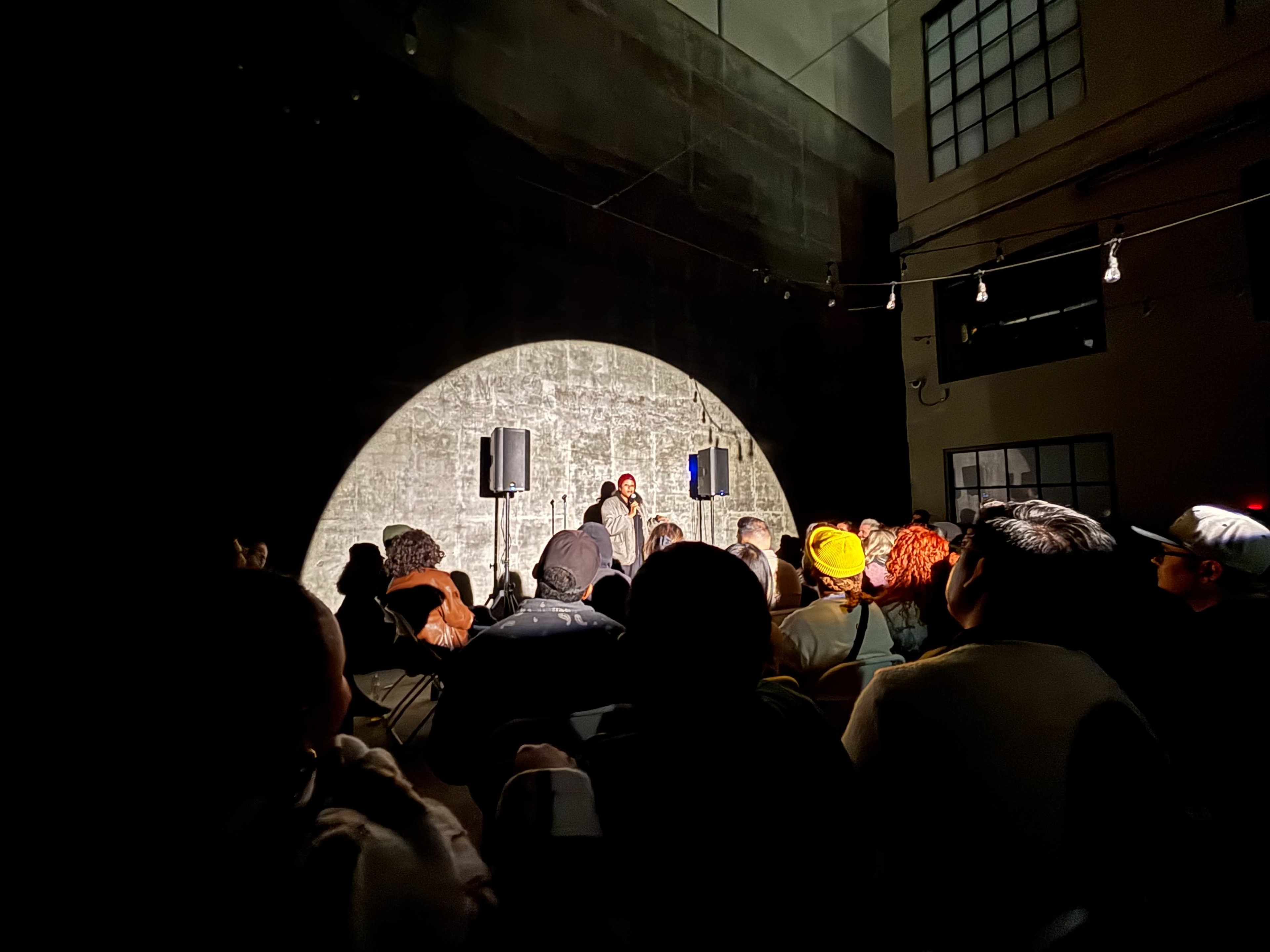 A performer stands on stage under a spotlight while an audience watches in a dimly lit venue.