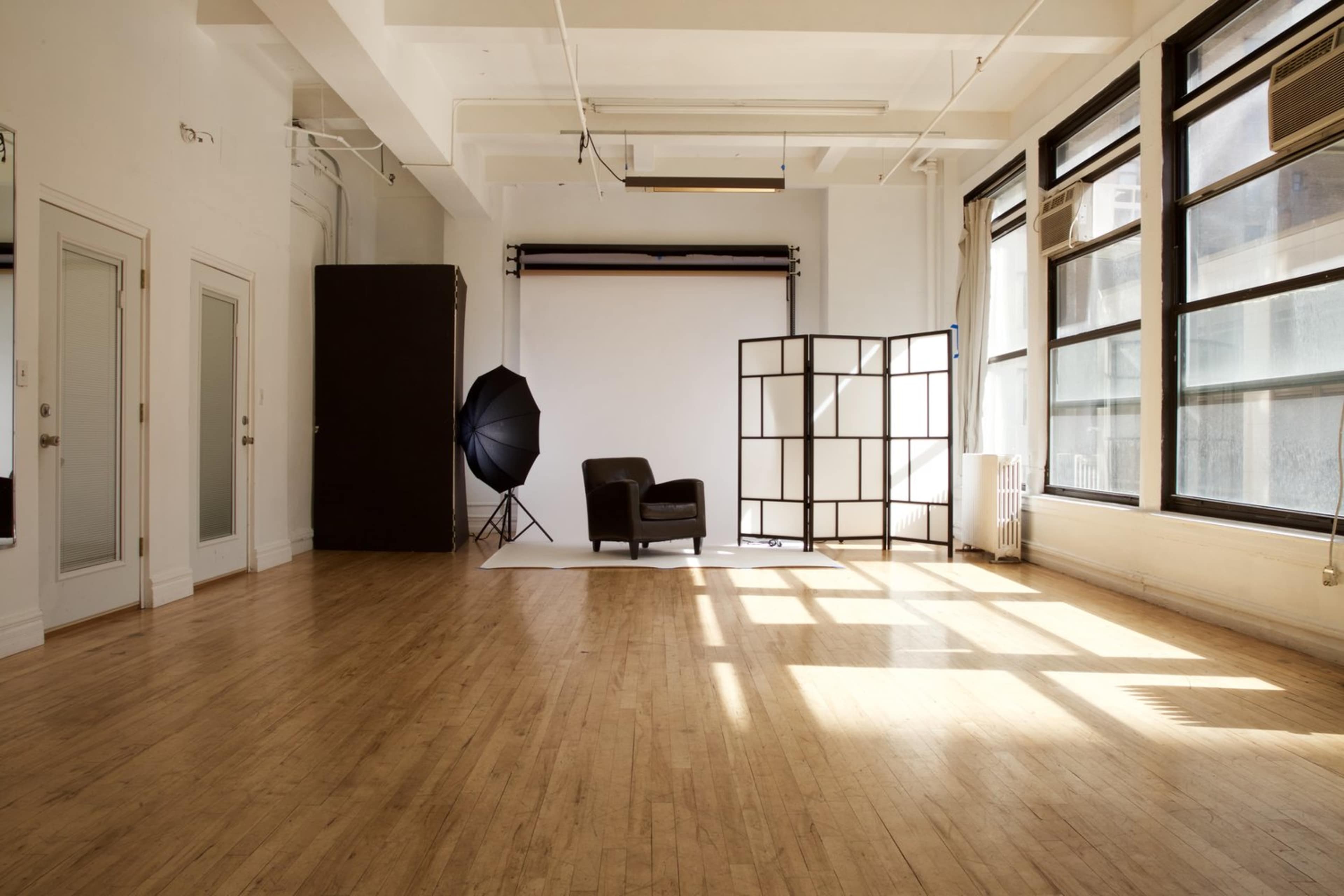 A bright, open studio space features a black chair, a backdrop setup, and large windows casting shadows on the wooden floor.