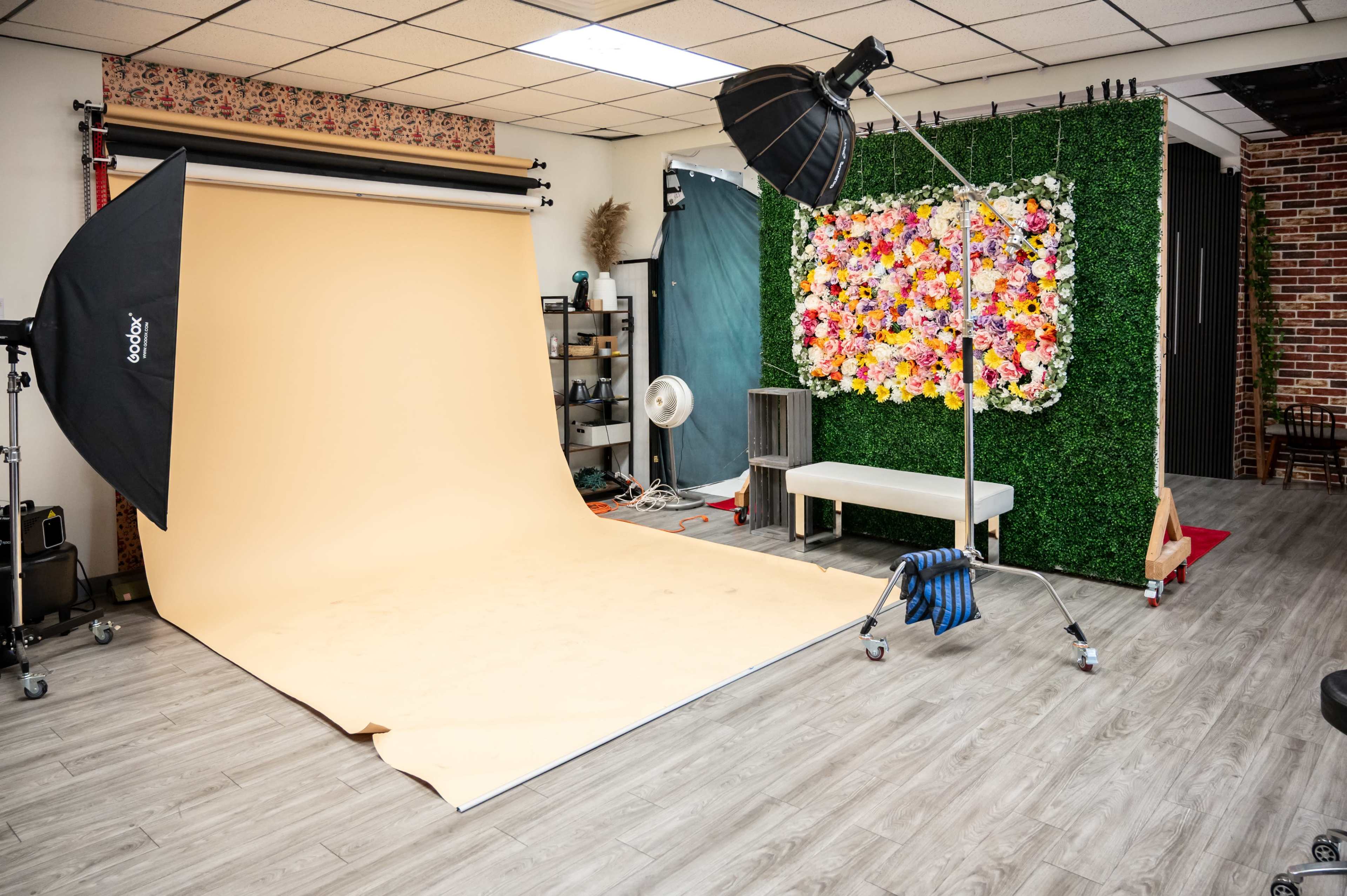 The image shows a photography studio with a beige backdrop, soft box lighting, a floral wall decoration, and various equipment arranged around the space.
