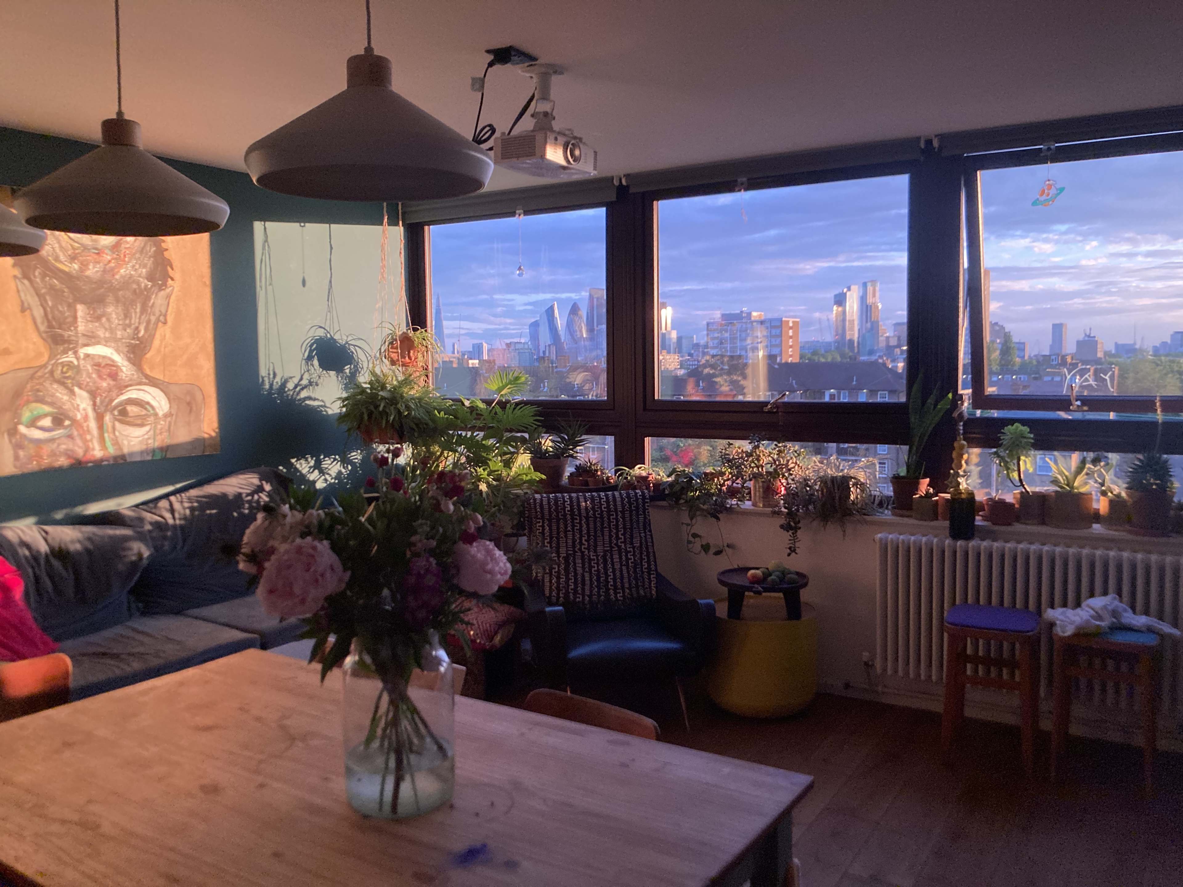 A cozy living room features a wooden table, a vase of flowers, and large windows showcasing a city skyline at sunset.