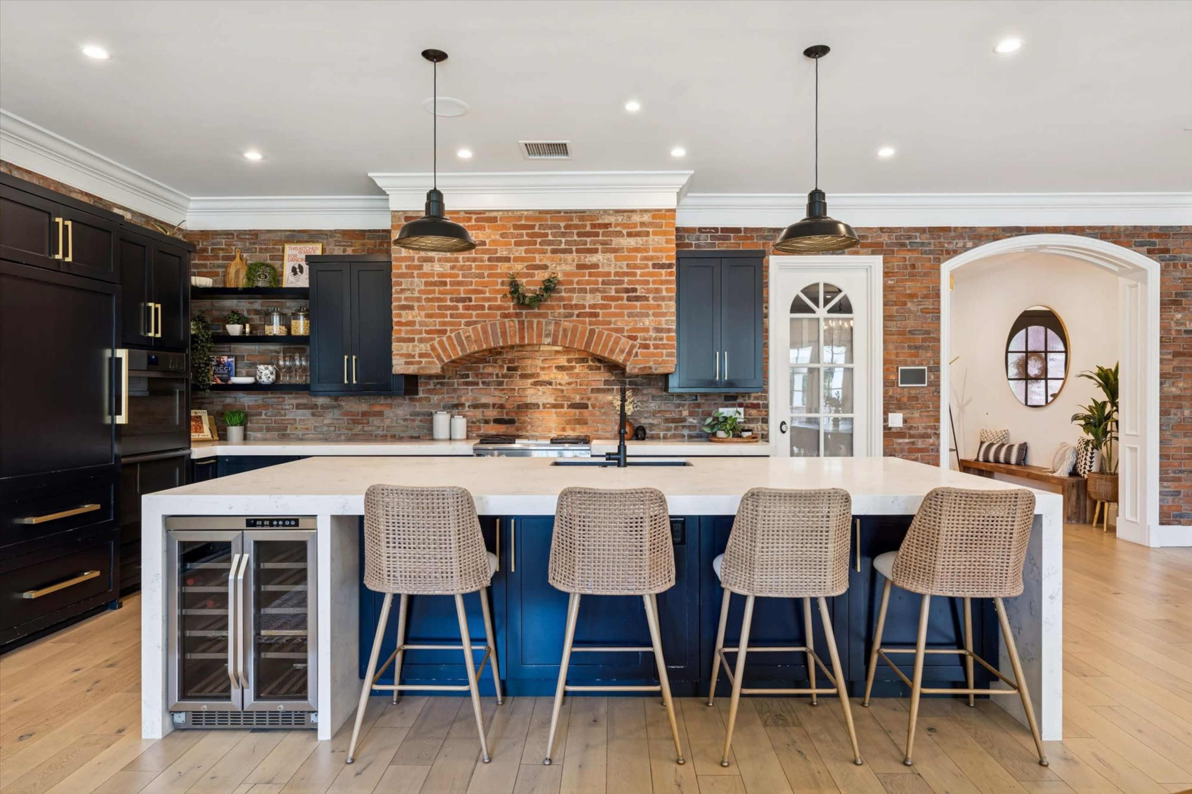 The kitchen features a spacious island with four wicker bar stools, a brick wall behind a stove, and dark cabinetry.