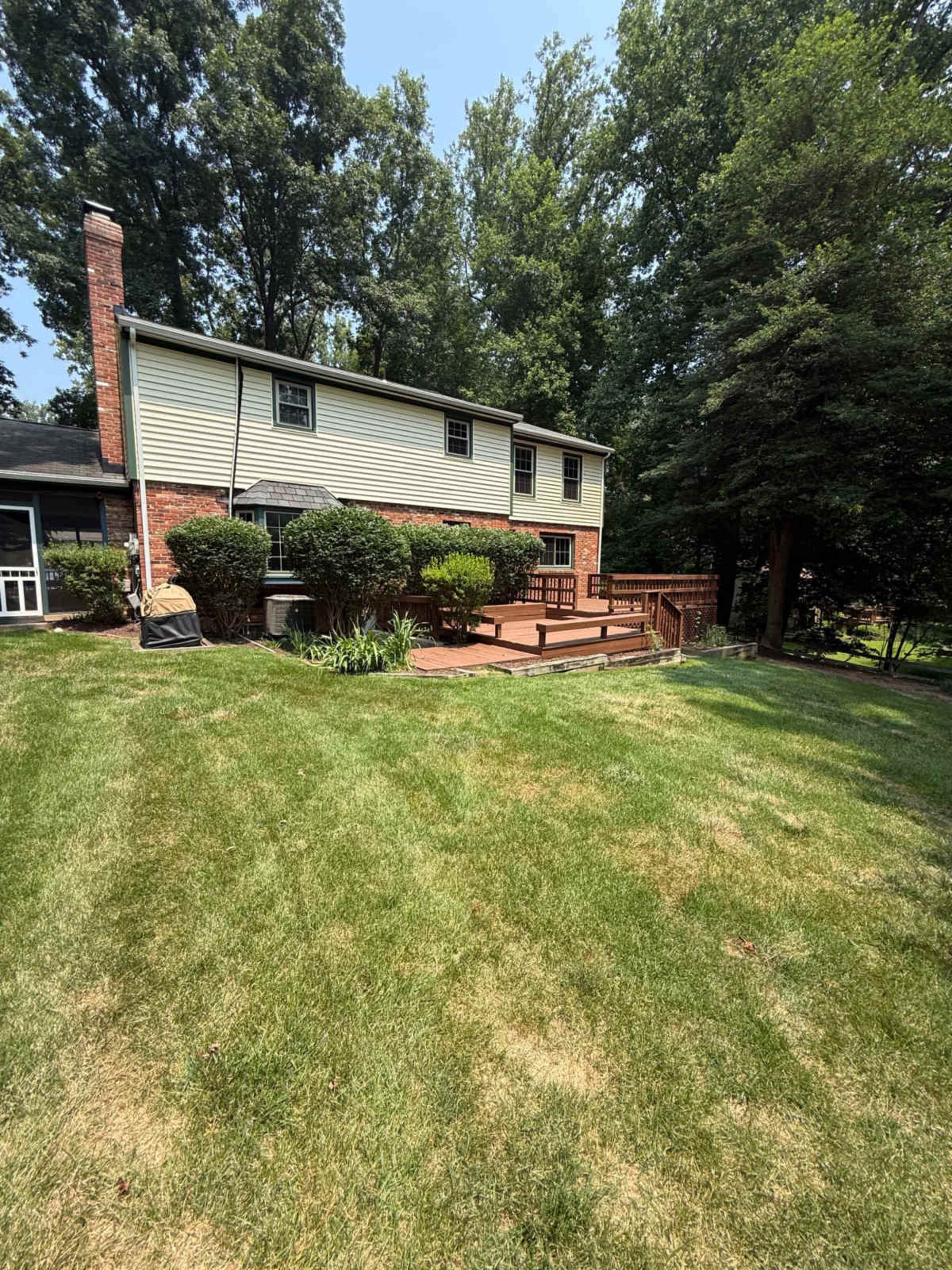 The image shows a two-story house with a wooden deck, surrounded by a manicured lawn and trees in the background.
