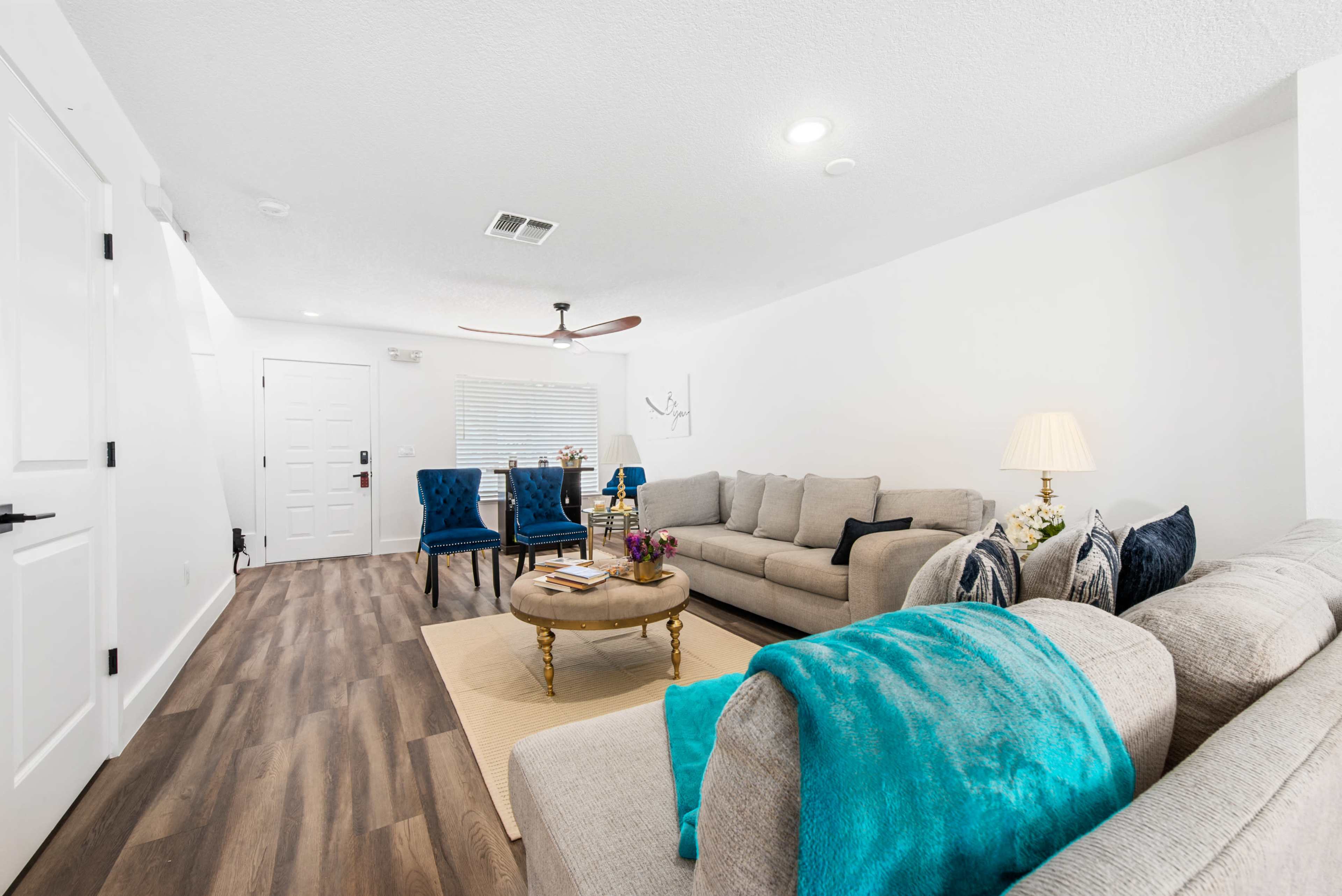 A bright living room features a light-colored sofa, a round coffee table with flowers, two blue armchairs, and a ceiling fan.