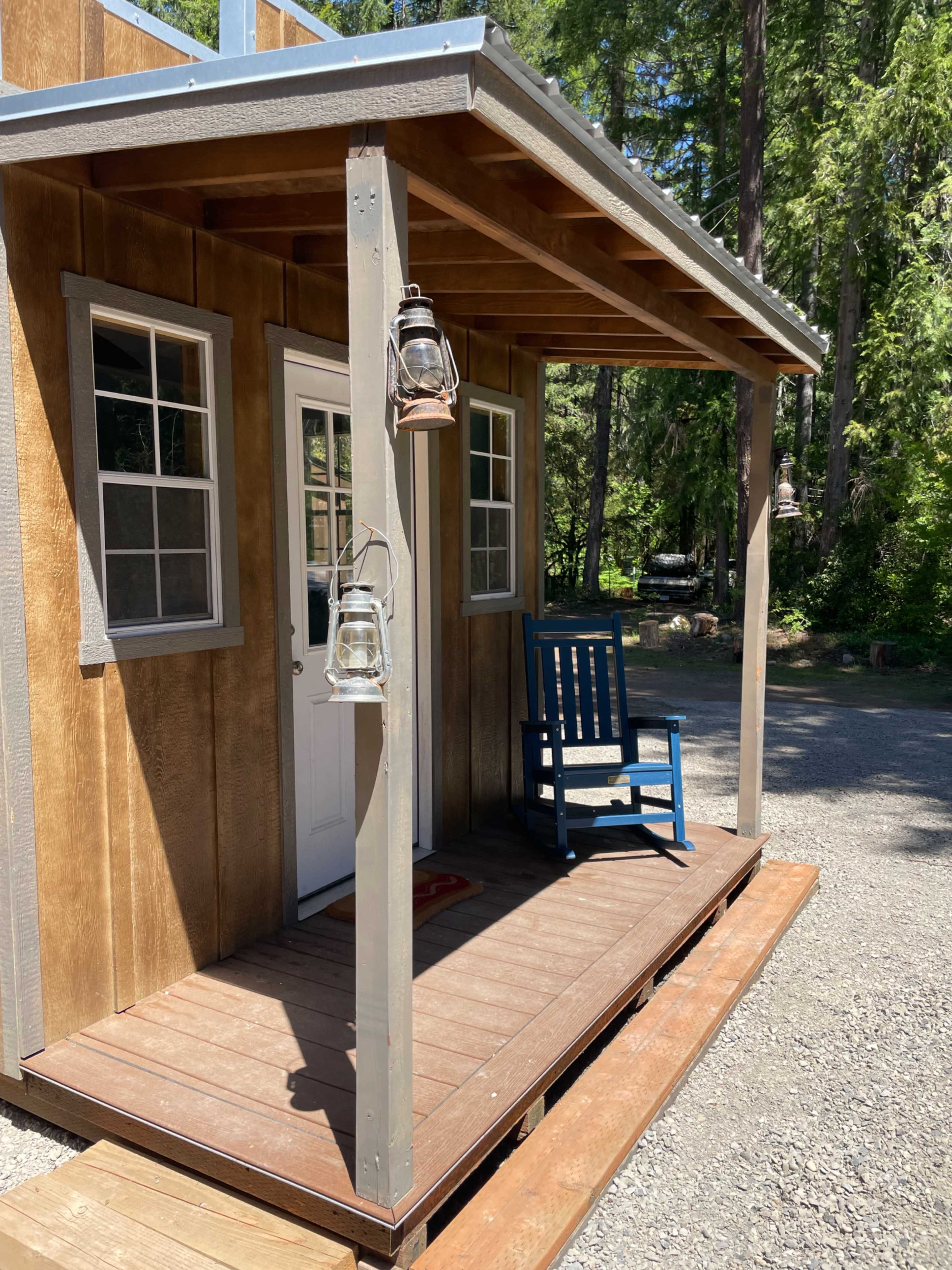 The image shows a small wooden cabin with a porch, featuring a blue rocking chair and two lanterns hanging beside the entrance.