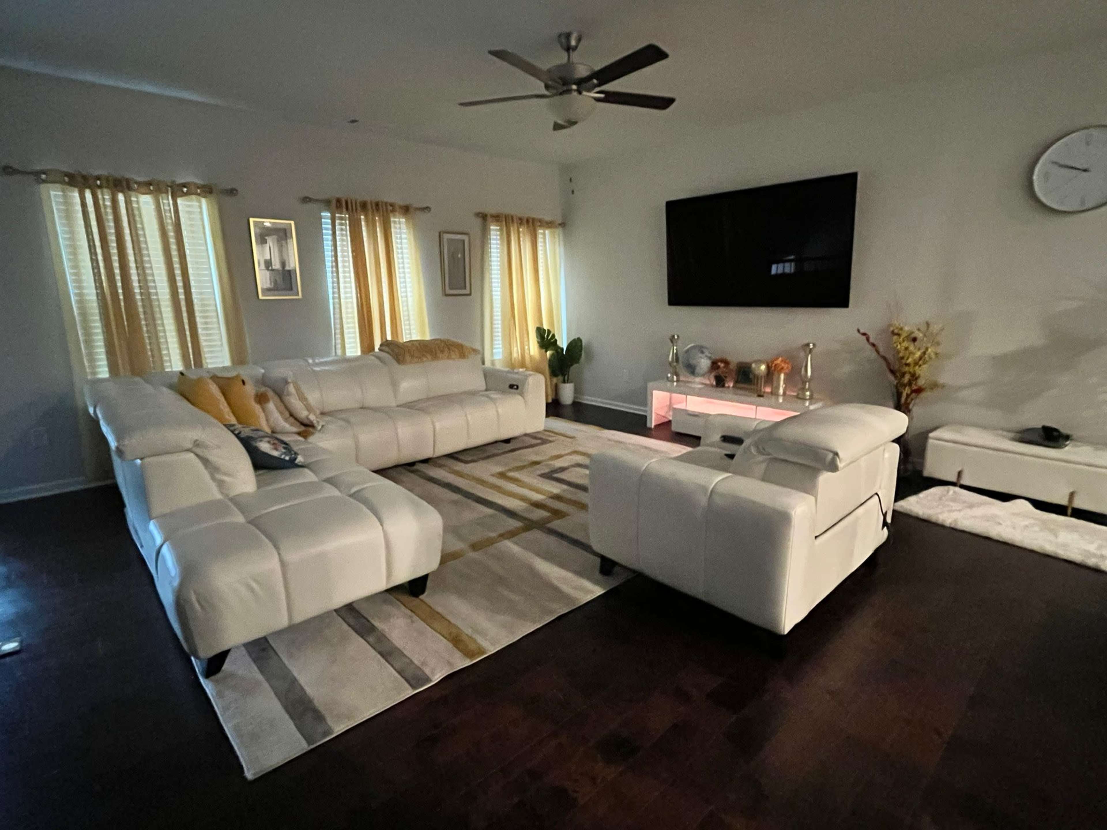 The image shows a modern living room with a white sectional sofa, a light-colored area rug, wooden flooring, and a large flat-screen TV mounted on the wall.