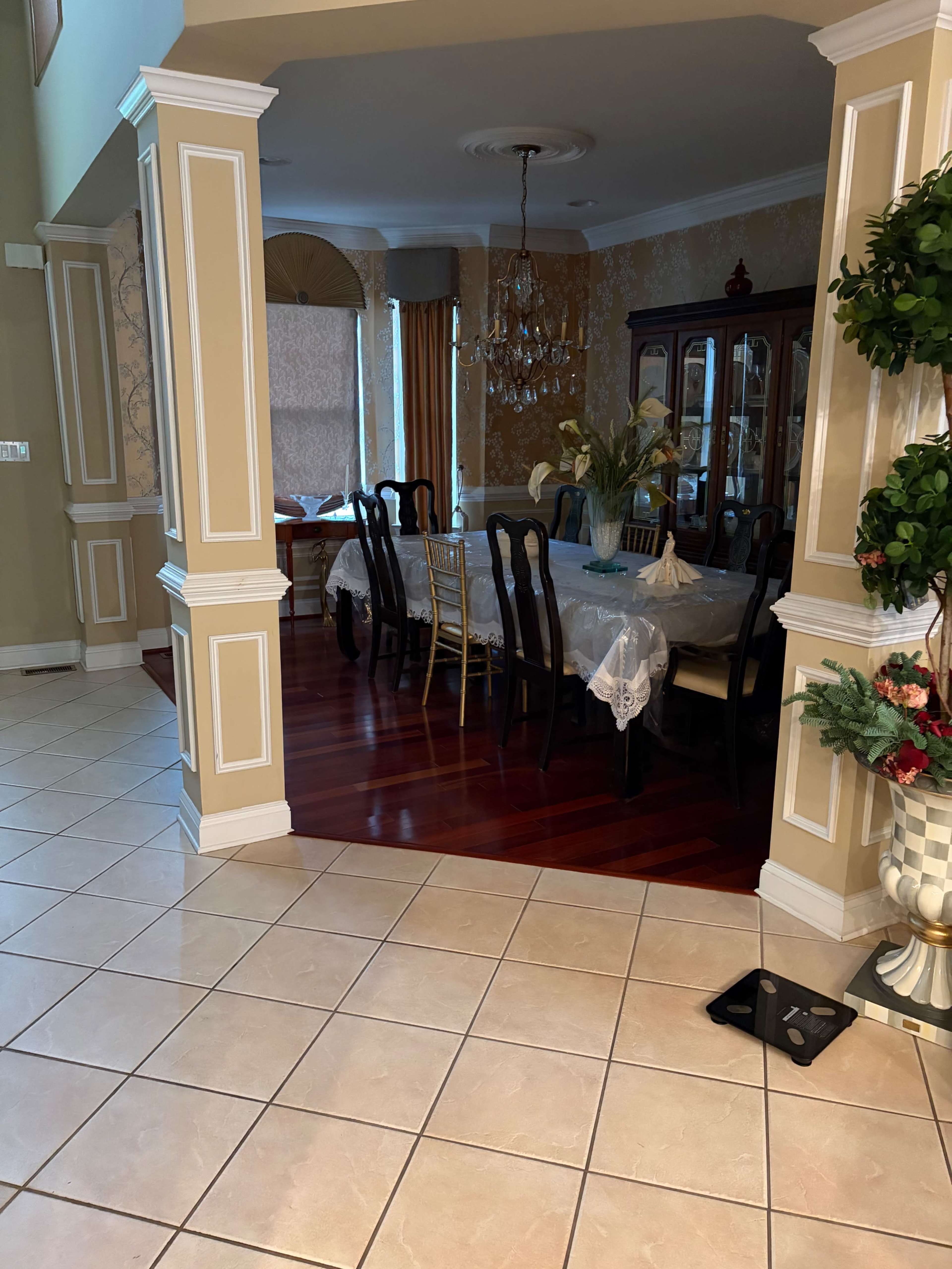 The image shows a dining area with a large table set with chairs, framed by decorative walls and a chandelier above.