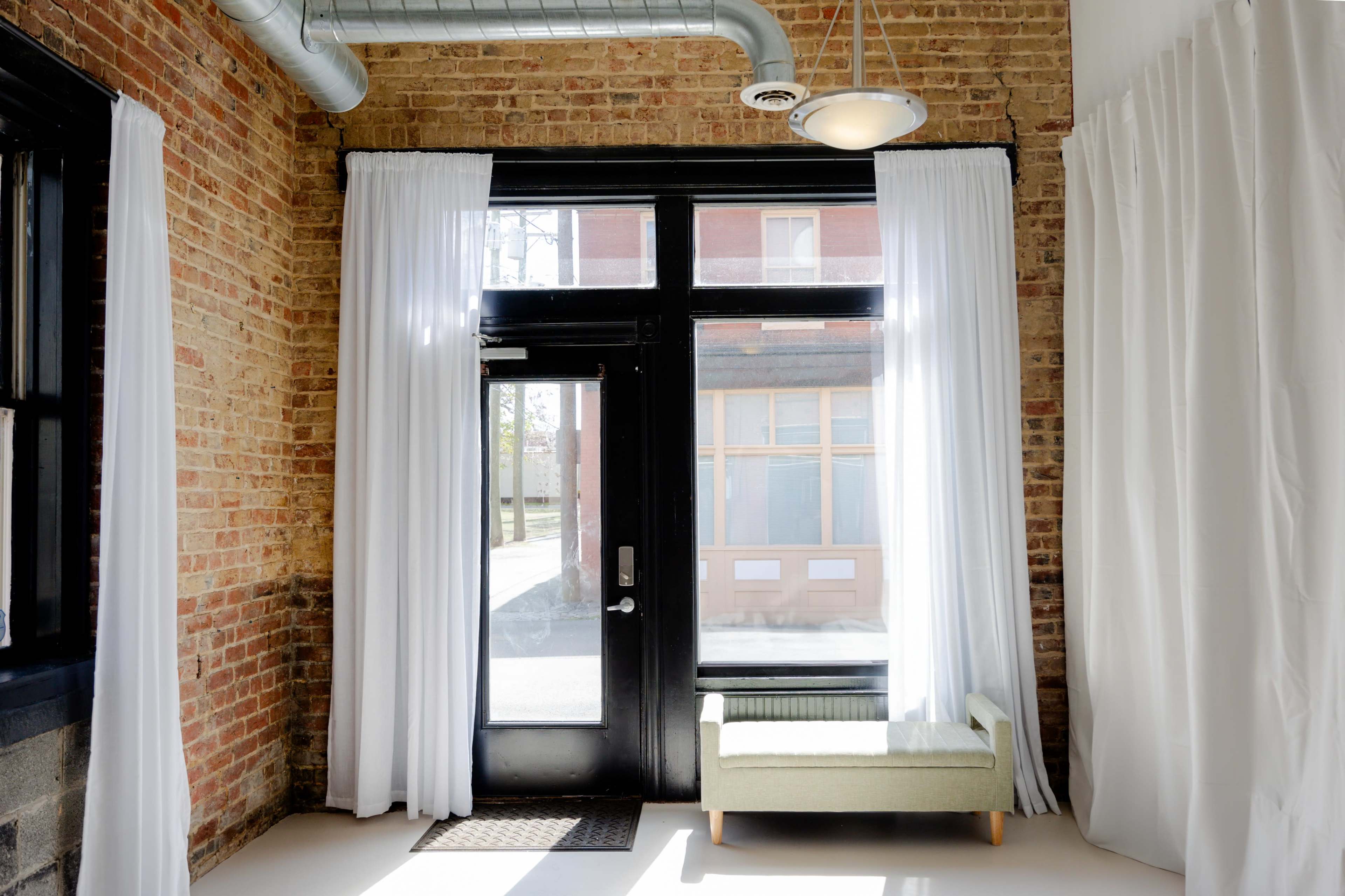 A room with a brick wall, large windows covered by white curtains, and a small green couch near the entrance.