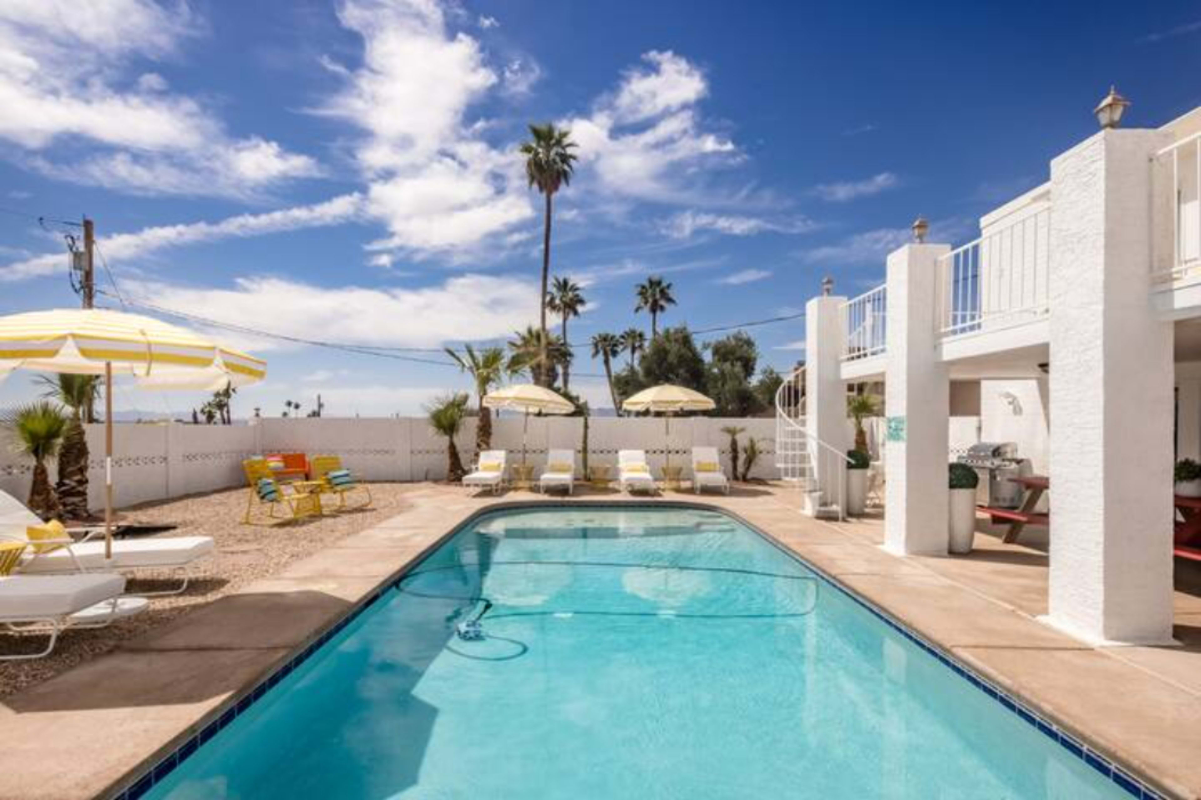 A sunny pool area with a clear blue pool, surrounded by lounge chairs and umbrellas, and palm trees in the background.