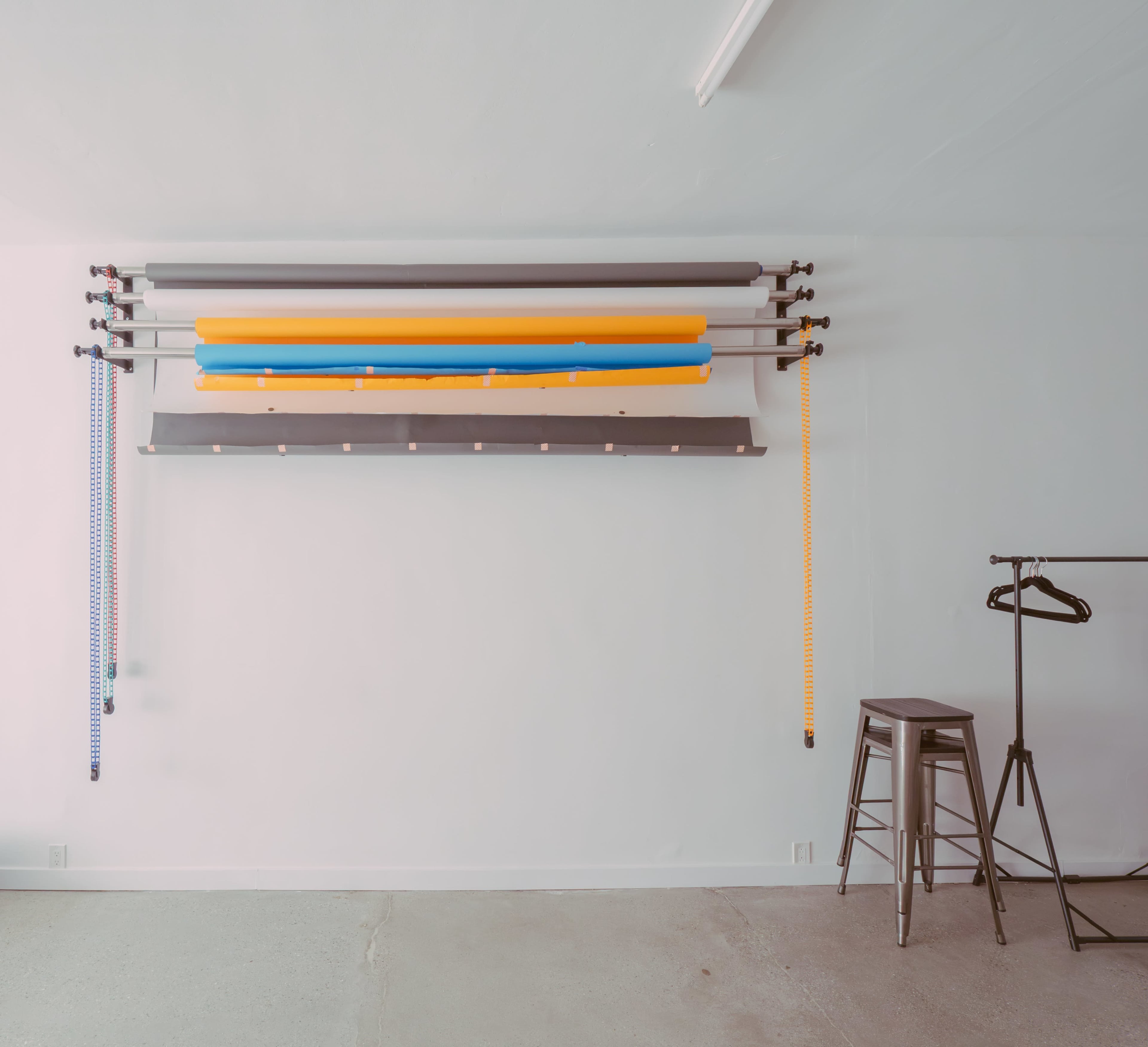 A wall-mounted rod system holds various colored rolls alongside a simple clothing rack and a stool in a minimalist room.