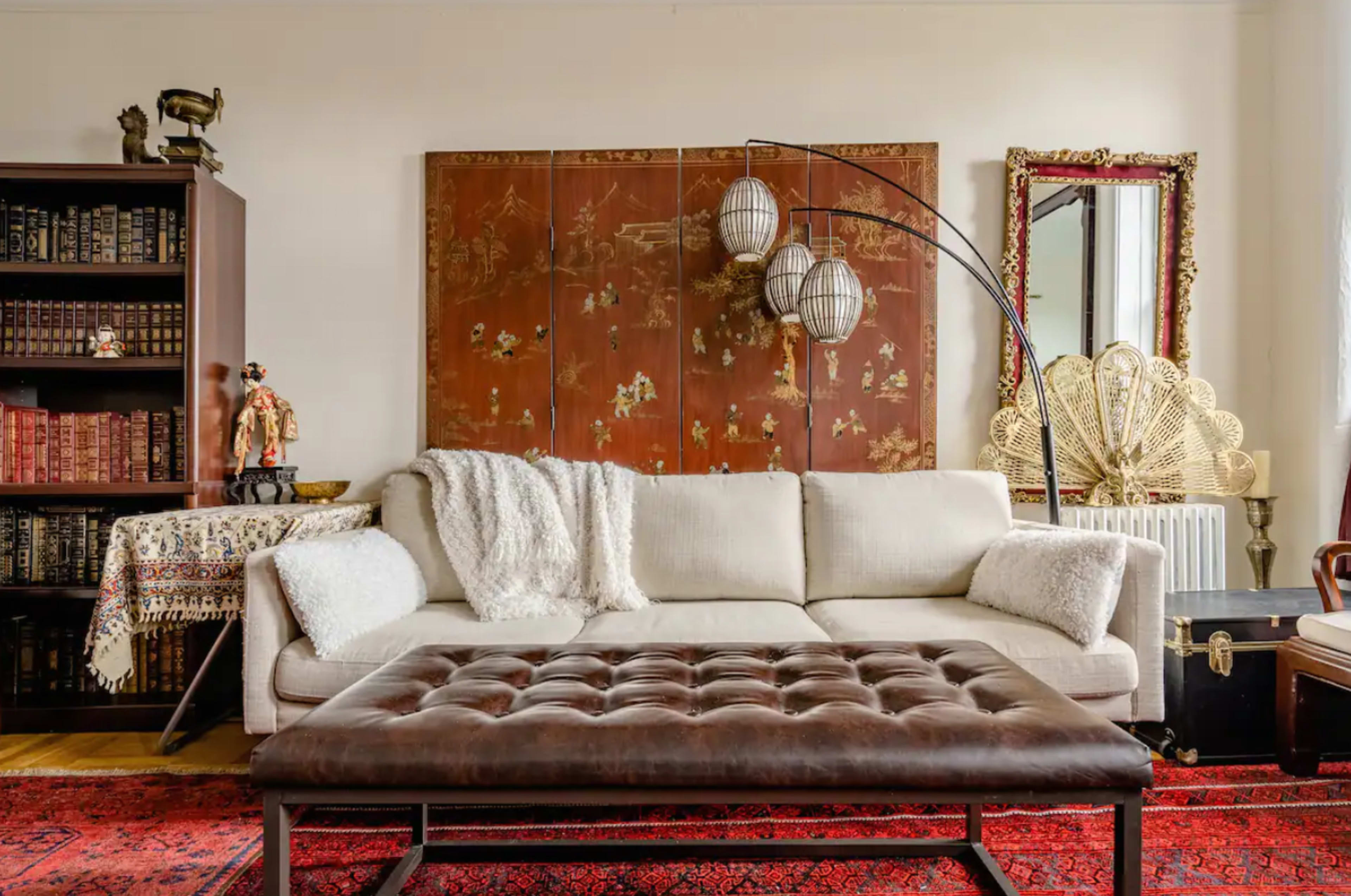 A living room with a white sofa, a brown tufted coffee table, a large decorative wall hanging, and various bookshelves filled with books.