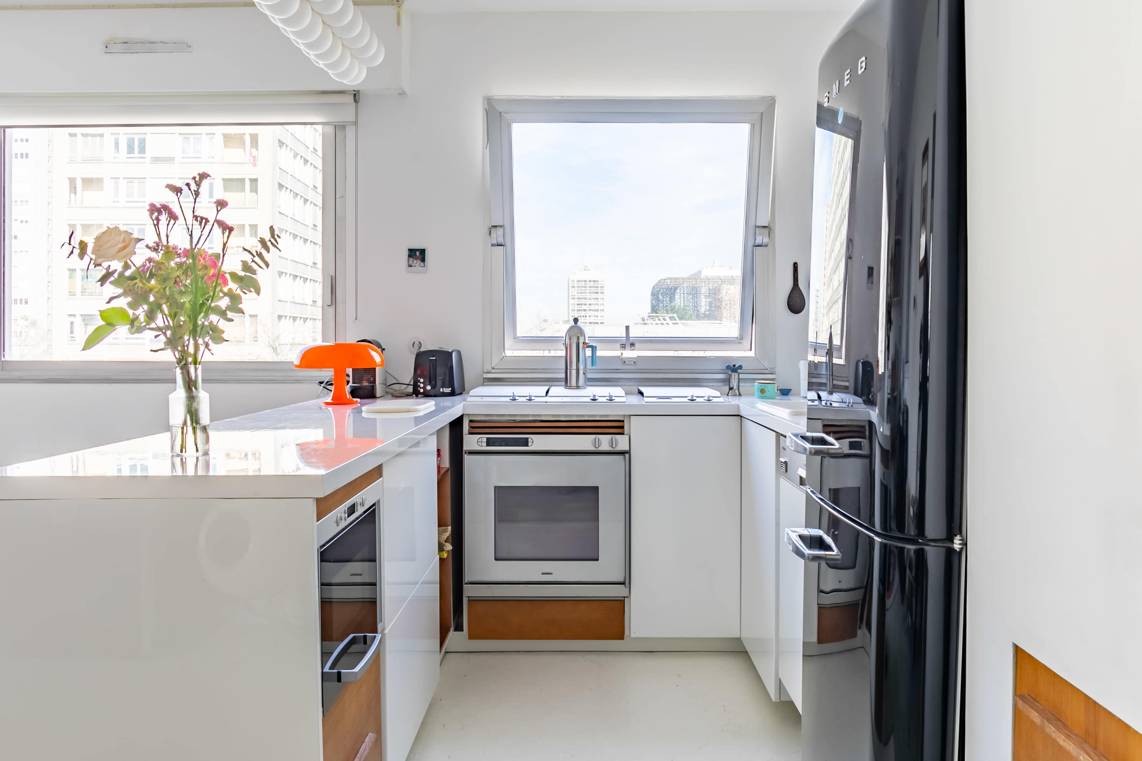 A modern kitchen features a black refrigerator, a white countertop, and large windows providing natural light and a view of the city.