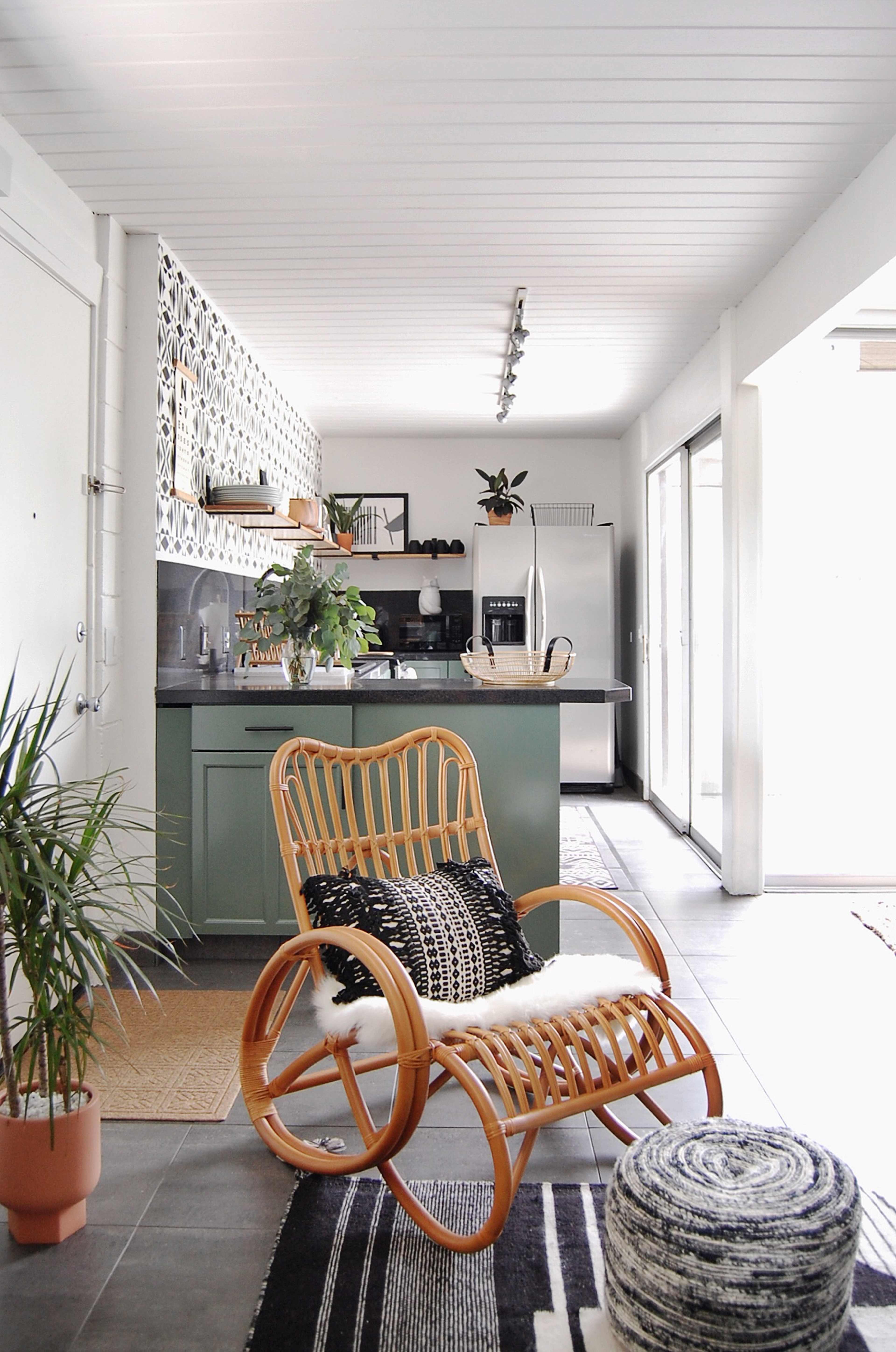 A wooden rocking chair with a cushion is positioned in a bright, modern room that leads to a kitchen featuring green cabinetry and patterned wall decor.