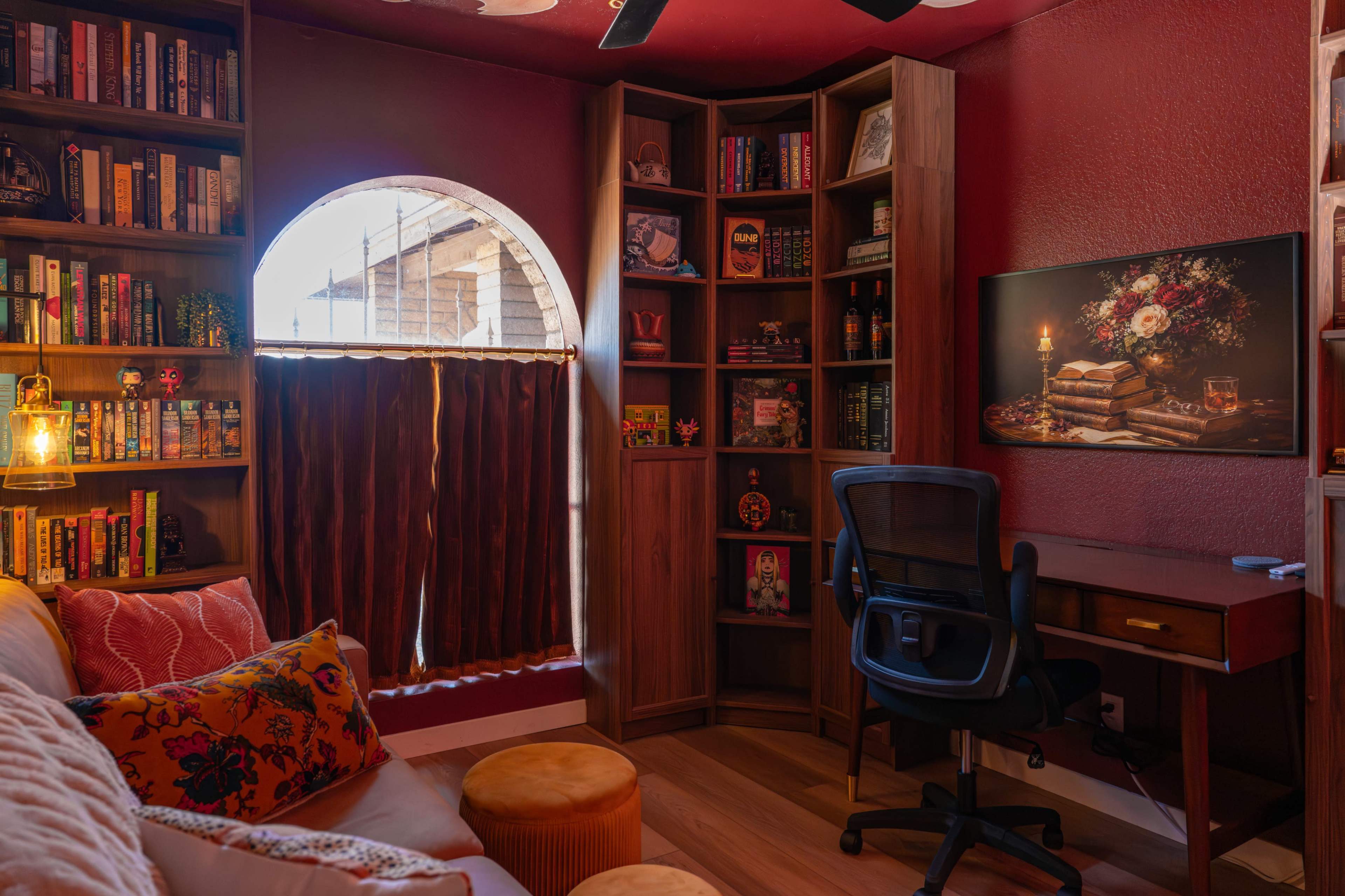 The image shows a cozy reading nook with a circular window, a plush sofa, wooden bookshelves filled with books, and a small desk with a chair.