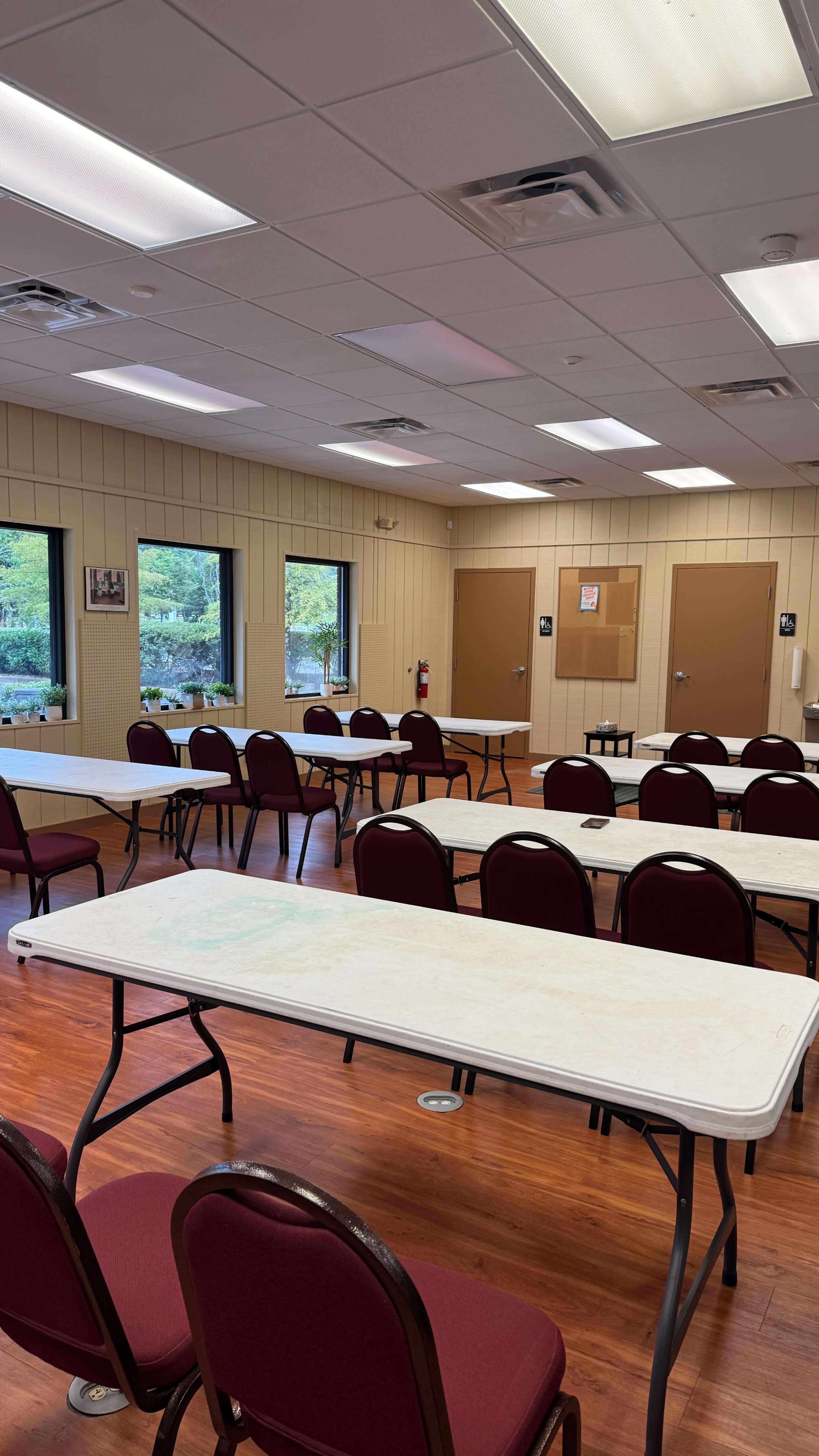 The image shows a sparsely furnished meeting room with several folding tables and chairs arranged in rows, along with windows letting in natural light.