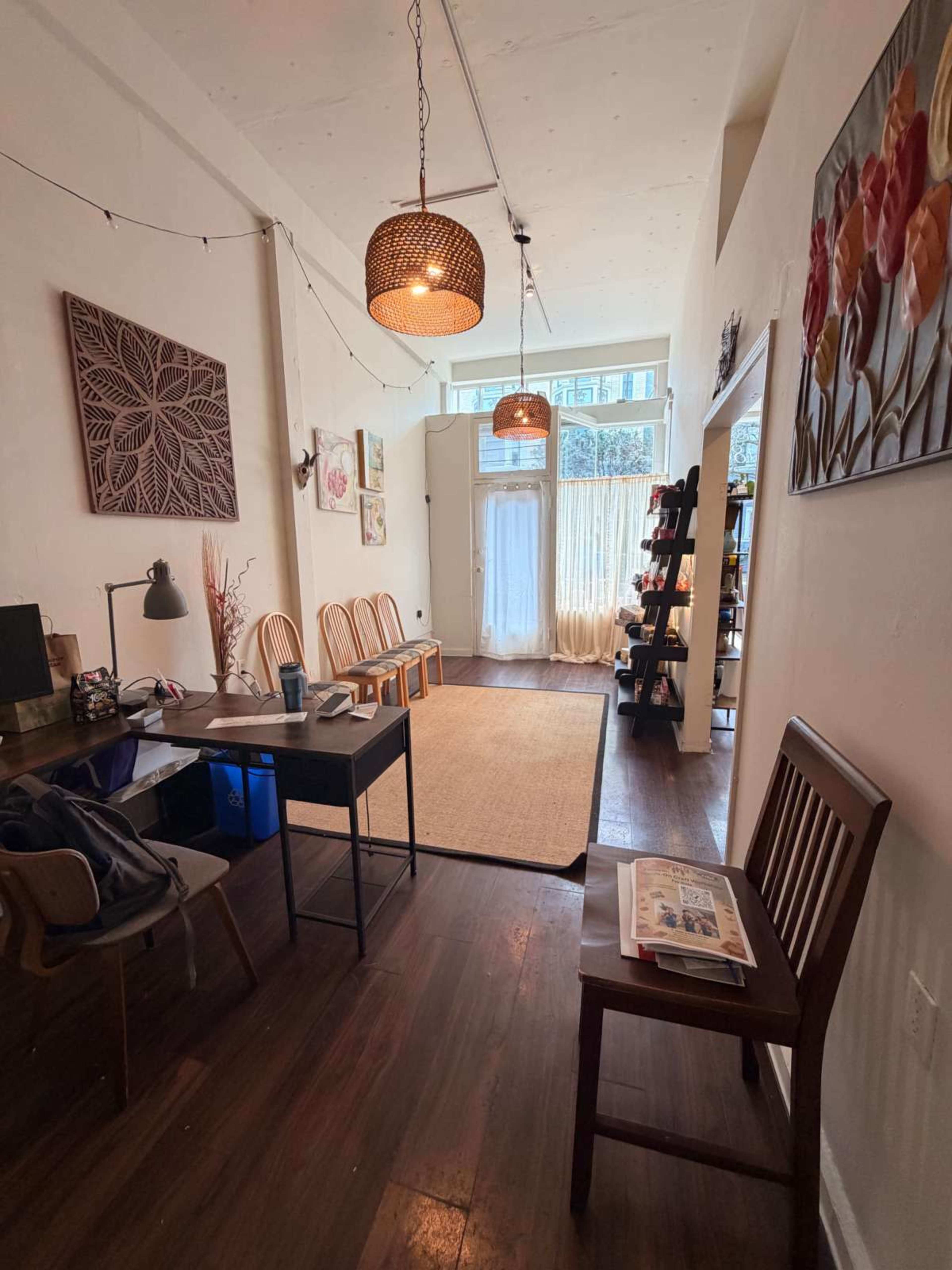 The image shows a cozy, minimalist room with wooden flooring, a table and chair set up on one side, and a bookshelf along the wall.