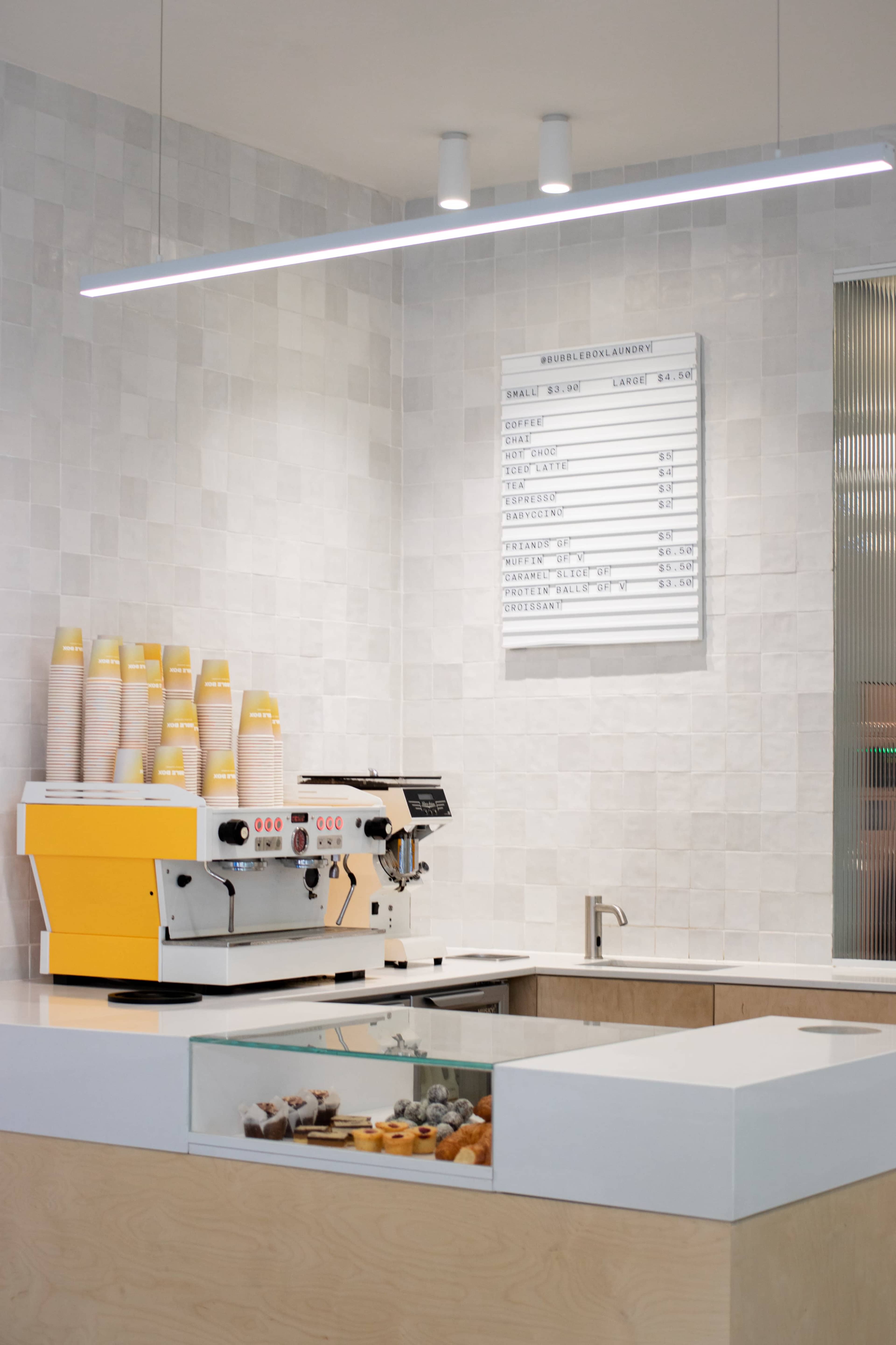 A modern café counter with a yellow espresso machine, stacked cups, a menu board, and a display case containing assorted pastries.