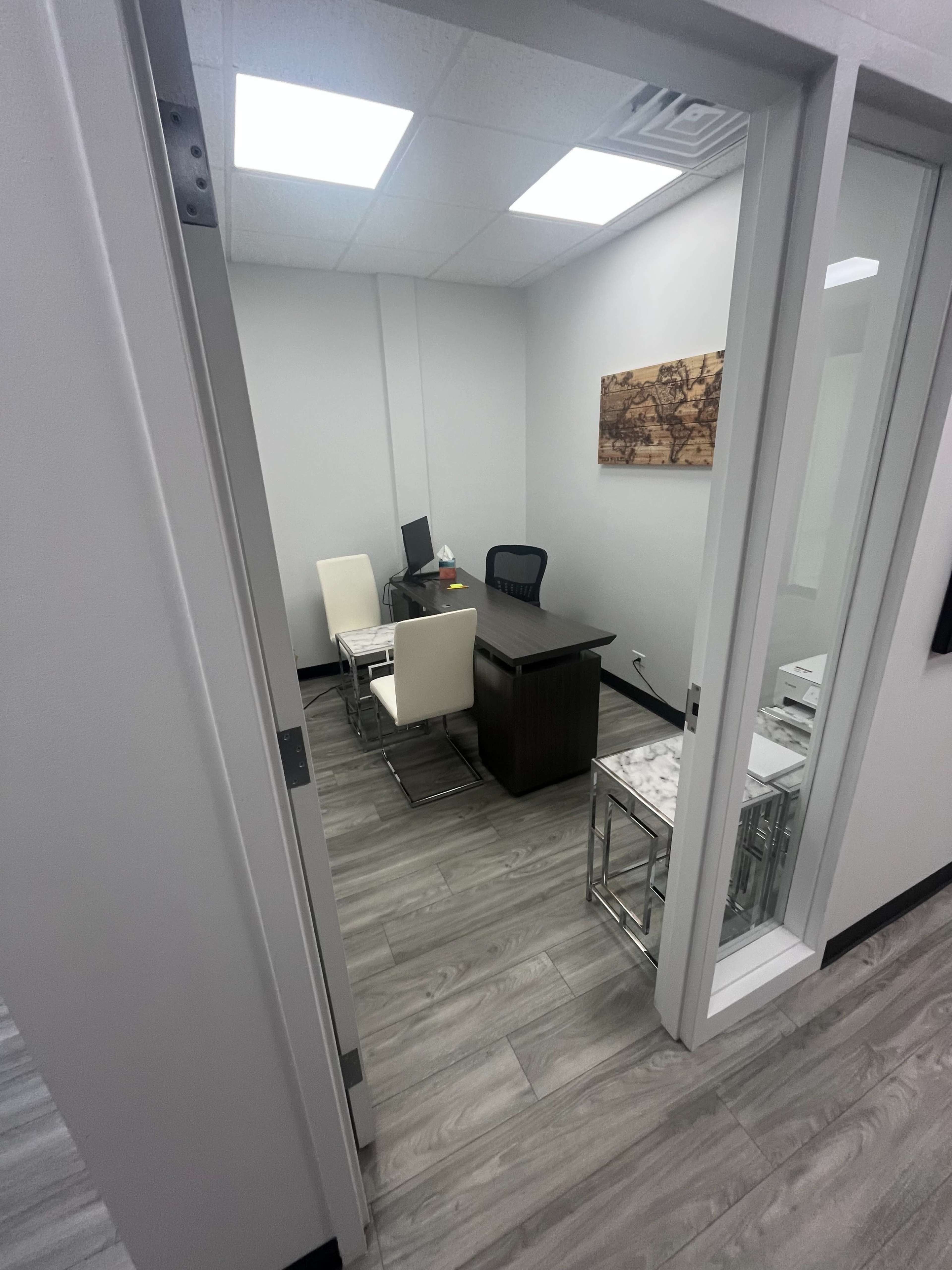The image shows a small office space with a desk, a chair, and several white chairs arranged around a table, all enclosed by glass walls.