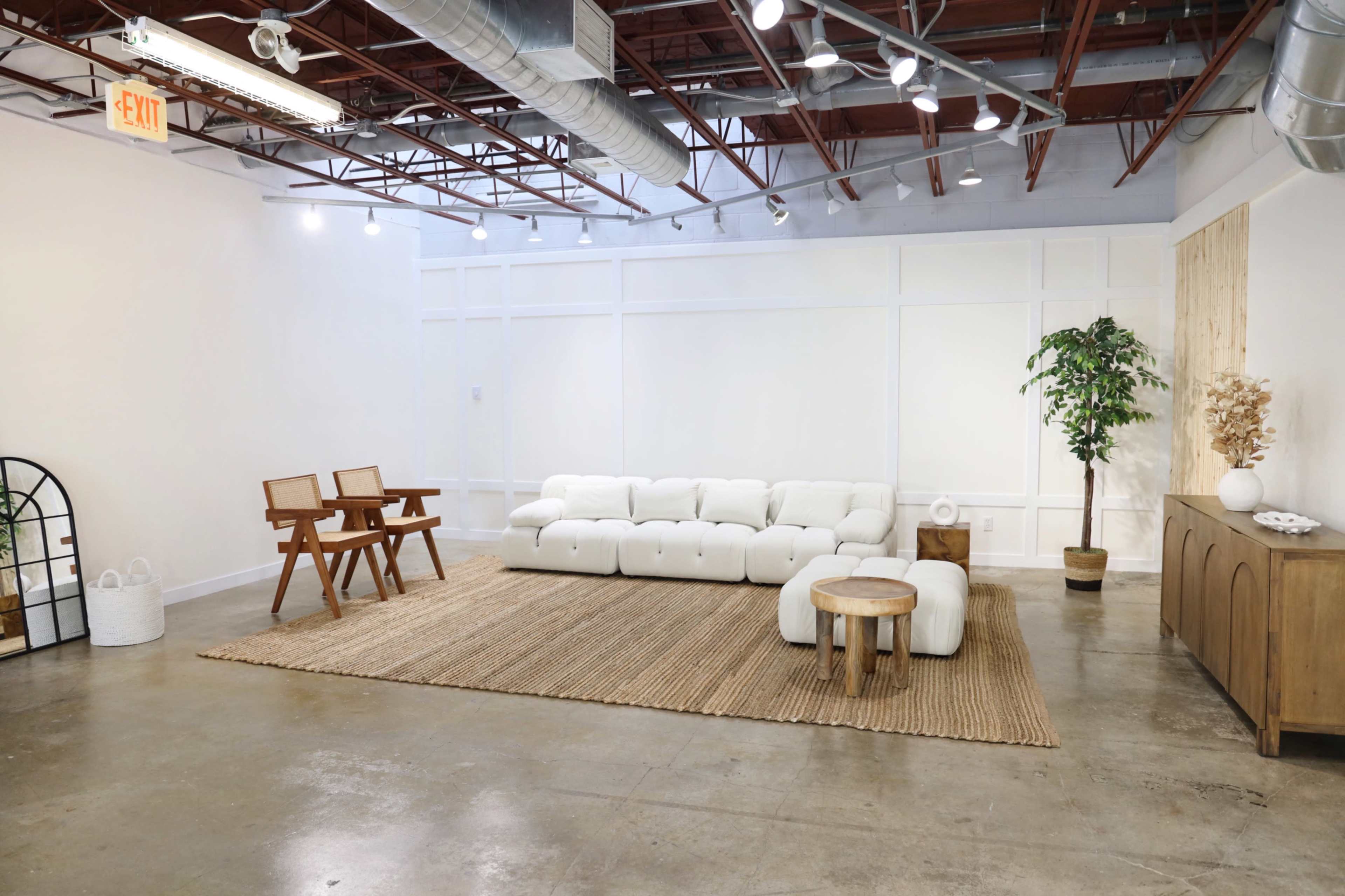 A spacious, minimalist interior features a white sectional sofa, two wooden chairs, a round wooden table, and a large area rug on polished concrete flooring.