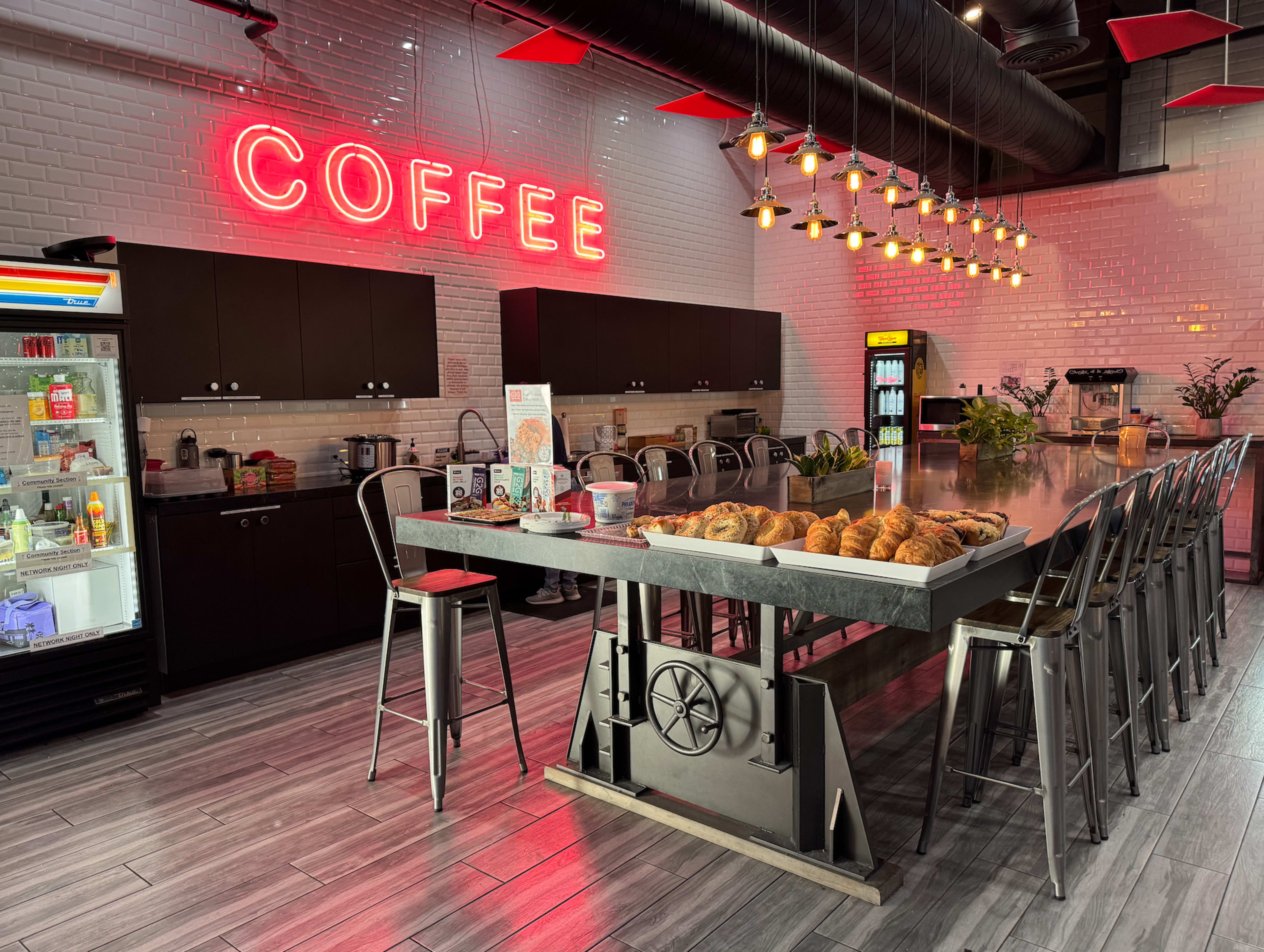 A modern coffee shop features a long countertop with metal stools, illuminated by a neon "COFFEE" sign and surrounded by various coffee equipment and pastries.