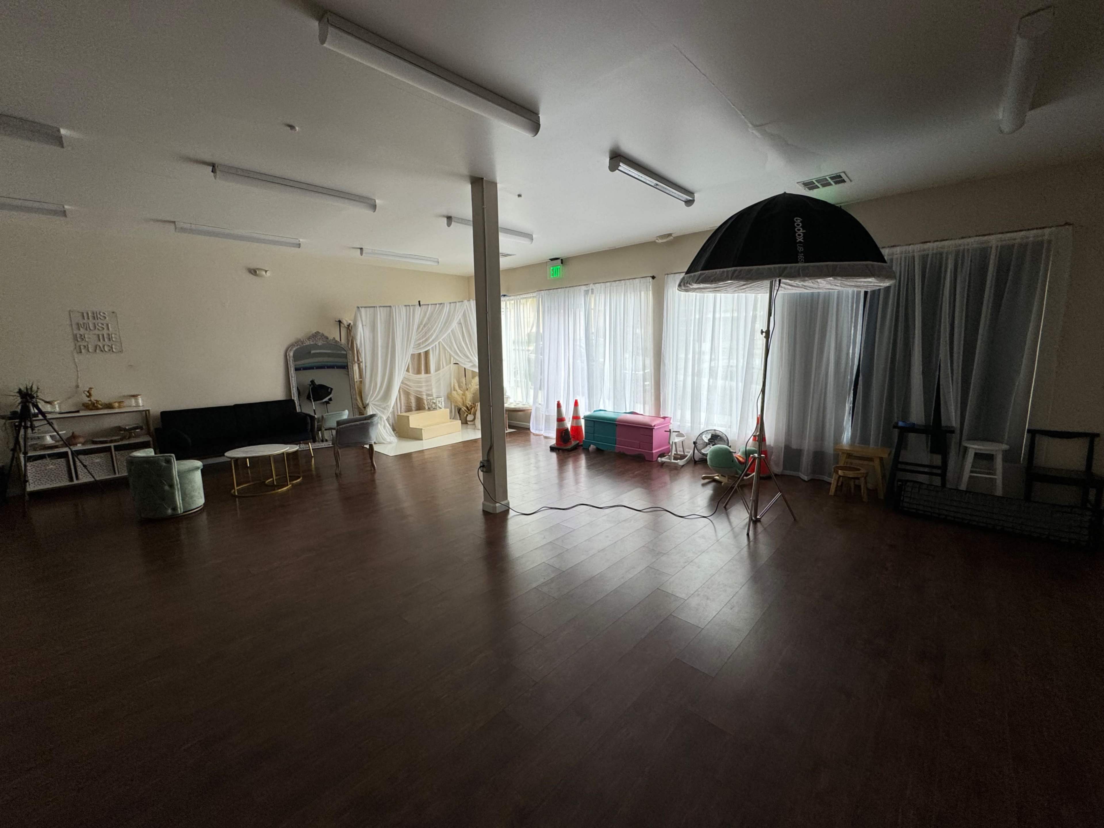 A spacious interior room with hardwood floors, large windows covered by sheer curtains, and various furniture pieces including a sofa, chairs, and a lighting setup.
