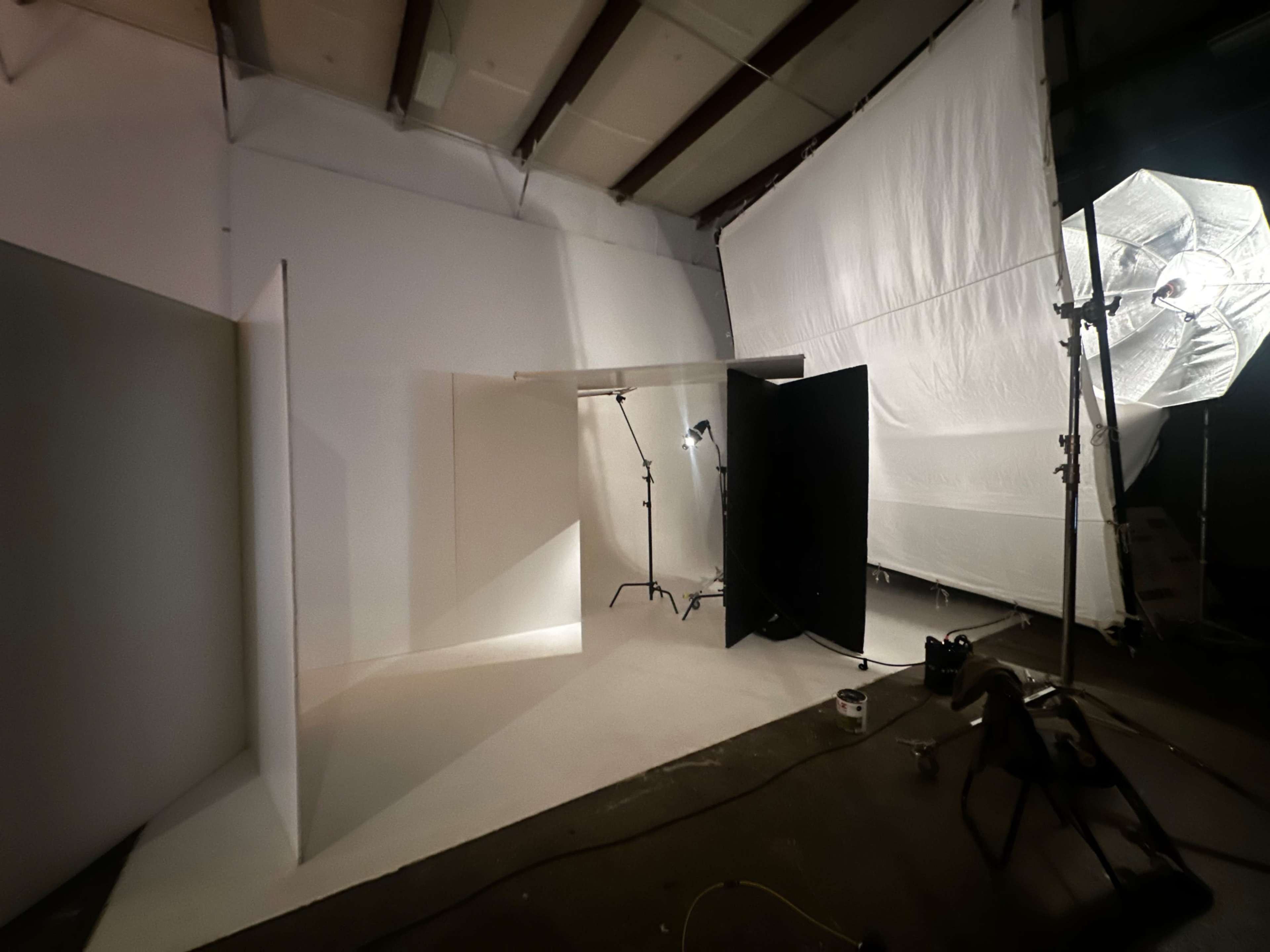 The image shows a photography studio setup with white backdrops, lighting equipment, and a black panel on a concrete floor.