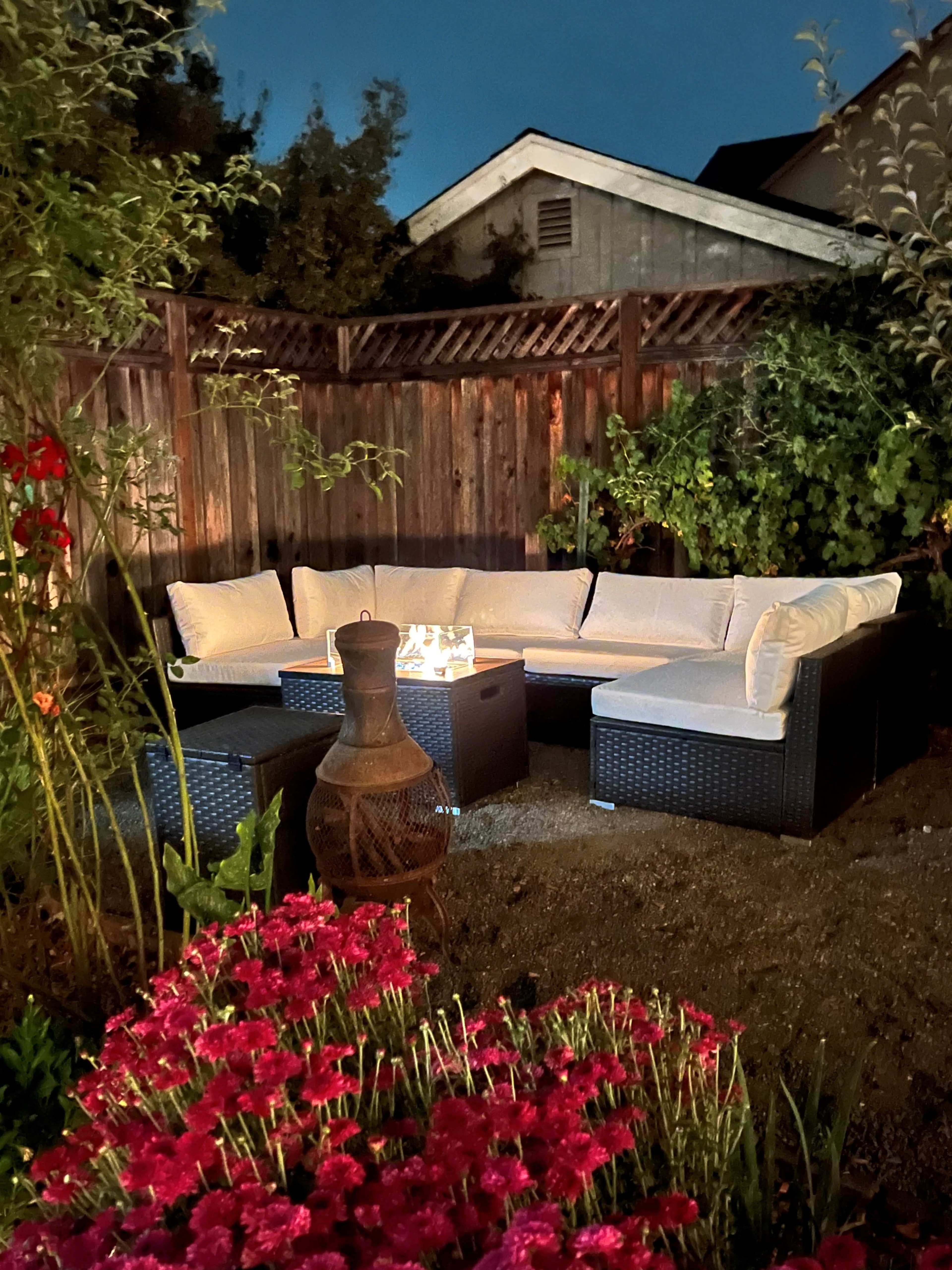 A well-lit outdoor seating area features a rectangular sectional sofa with cushions, surrounded by flowers and illuminated by soft lighting.