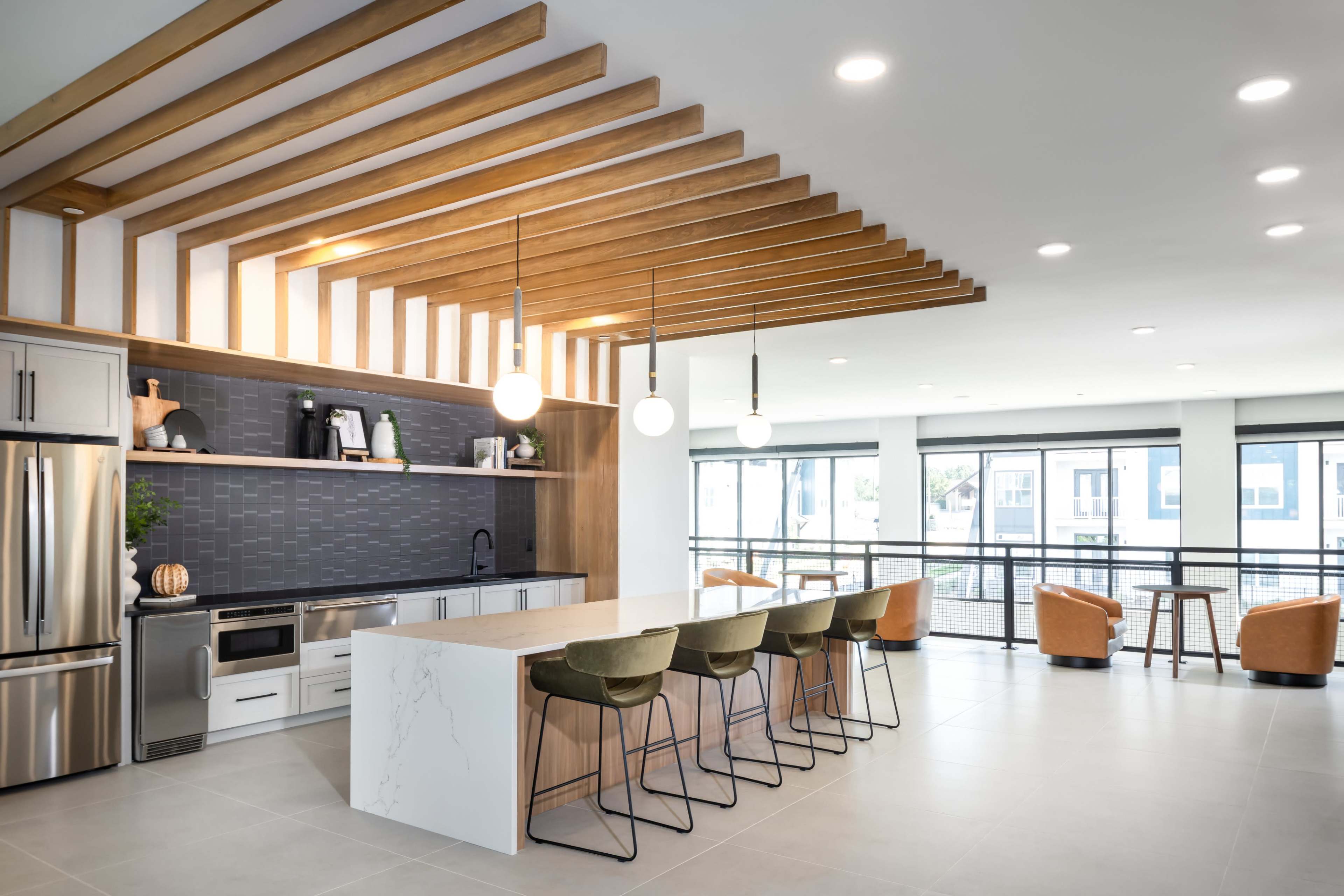 The image shows a modern kitchen and dining area with a large island, bar stools, a sleek countertop, and large windows allowing natural light to enter.