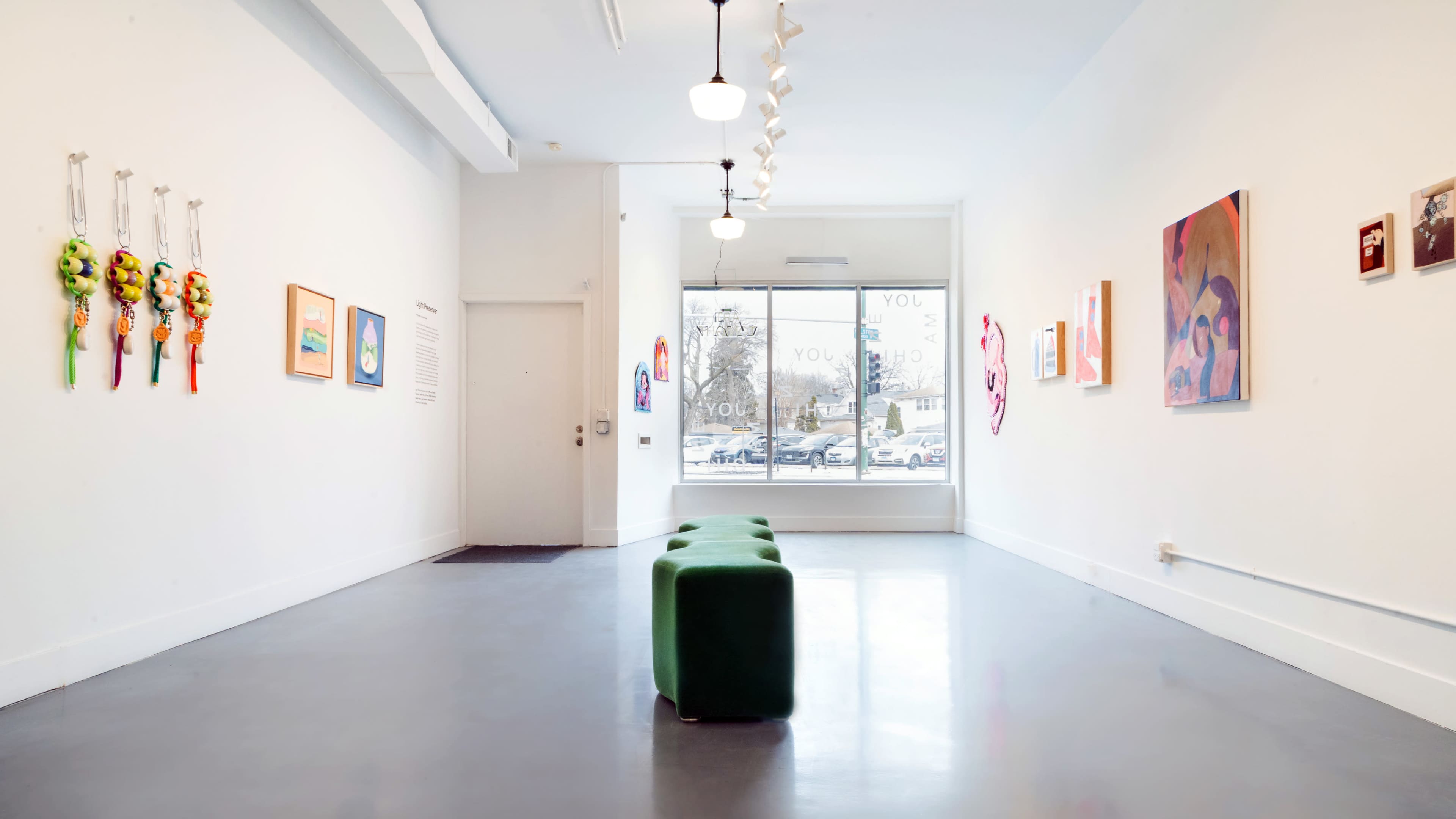 The image shows a modern art gallery with white walls, featuring various artworks hung on the walls and a green bench positioned in the center.