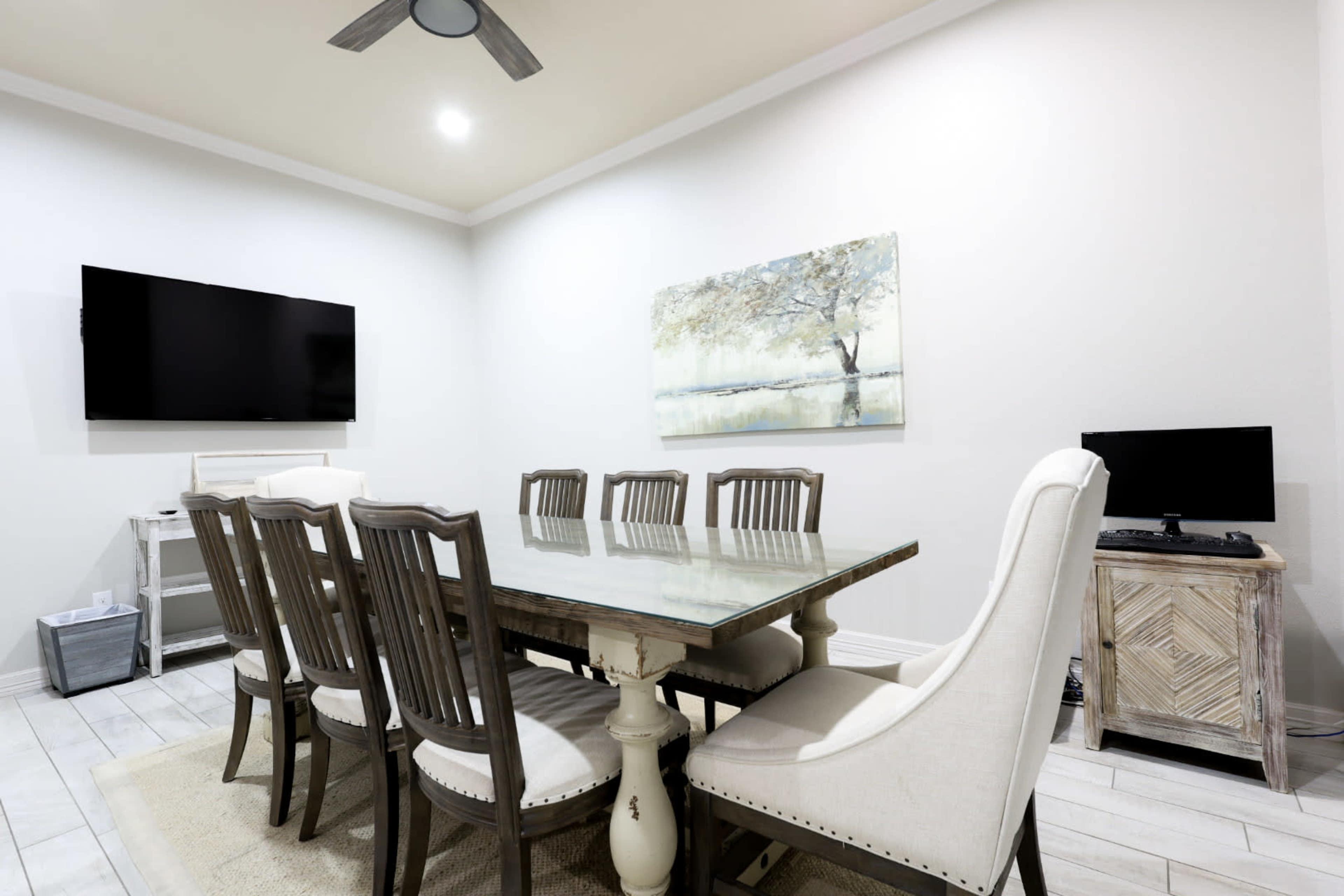 A modern dining room with a large table surrounded by eight chairs, a wall-mounted TV, and a framed painting of a tree on the wall.