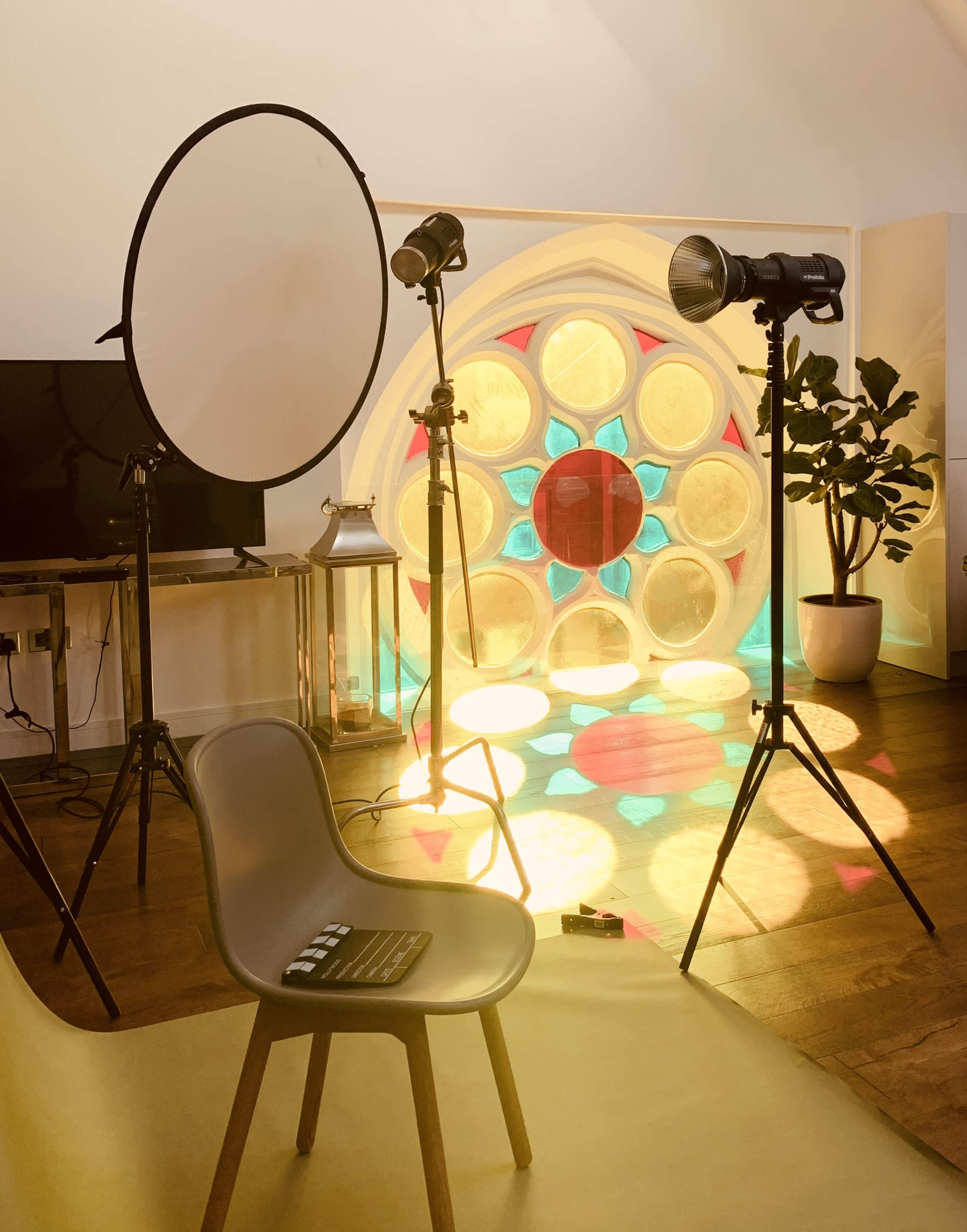 A photography setup featuring two lights, a large circular reflector, a chair, and a colorful stained glass window in a well-lit interior space.