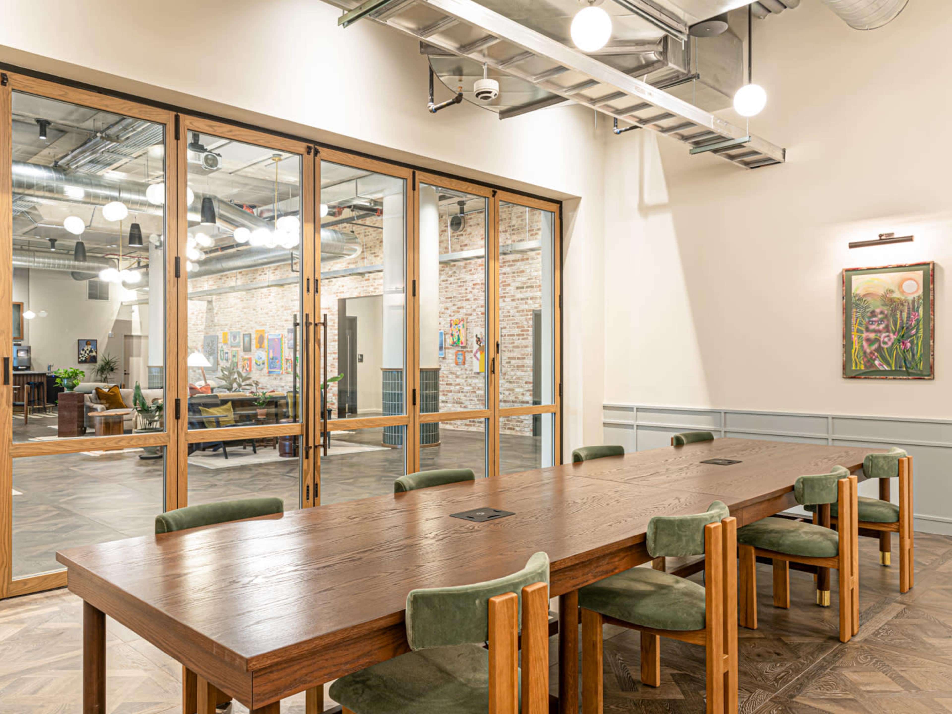 A large wooden table with green chairs in a well-lit room, featuring glass doors that lead to another space decorated with art and plants.