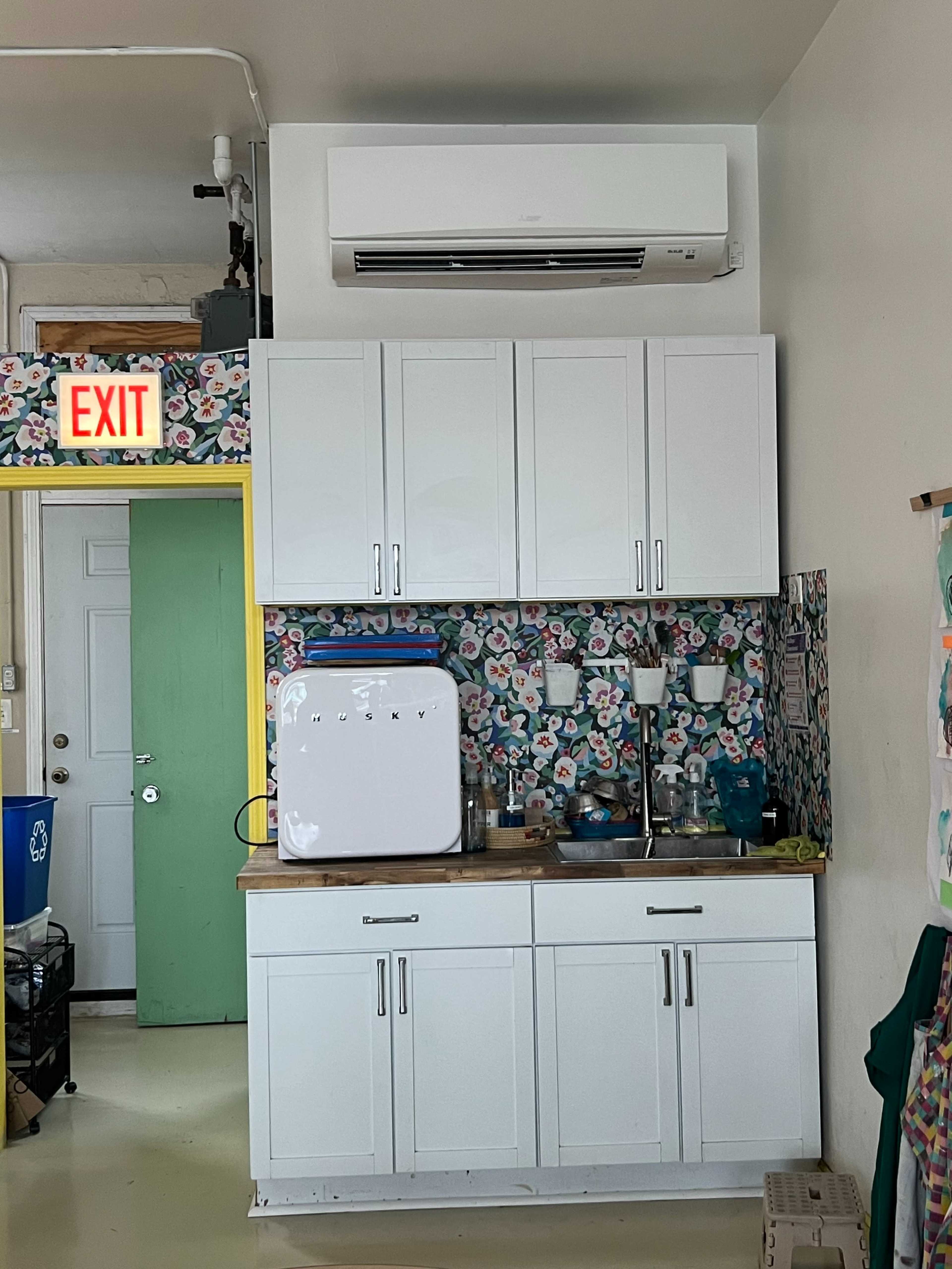 The image shows a small kitchen area with a white refrigerator, cabinets above a countertop, and a colorful floral wallpaper in the background, along with an exit sign and a green door.