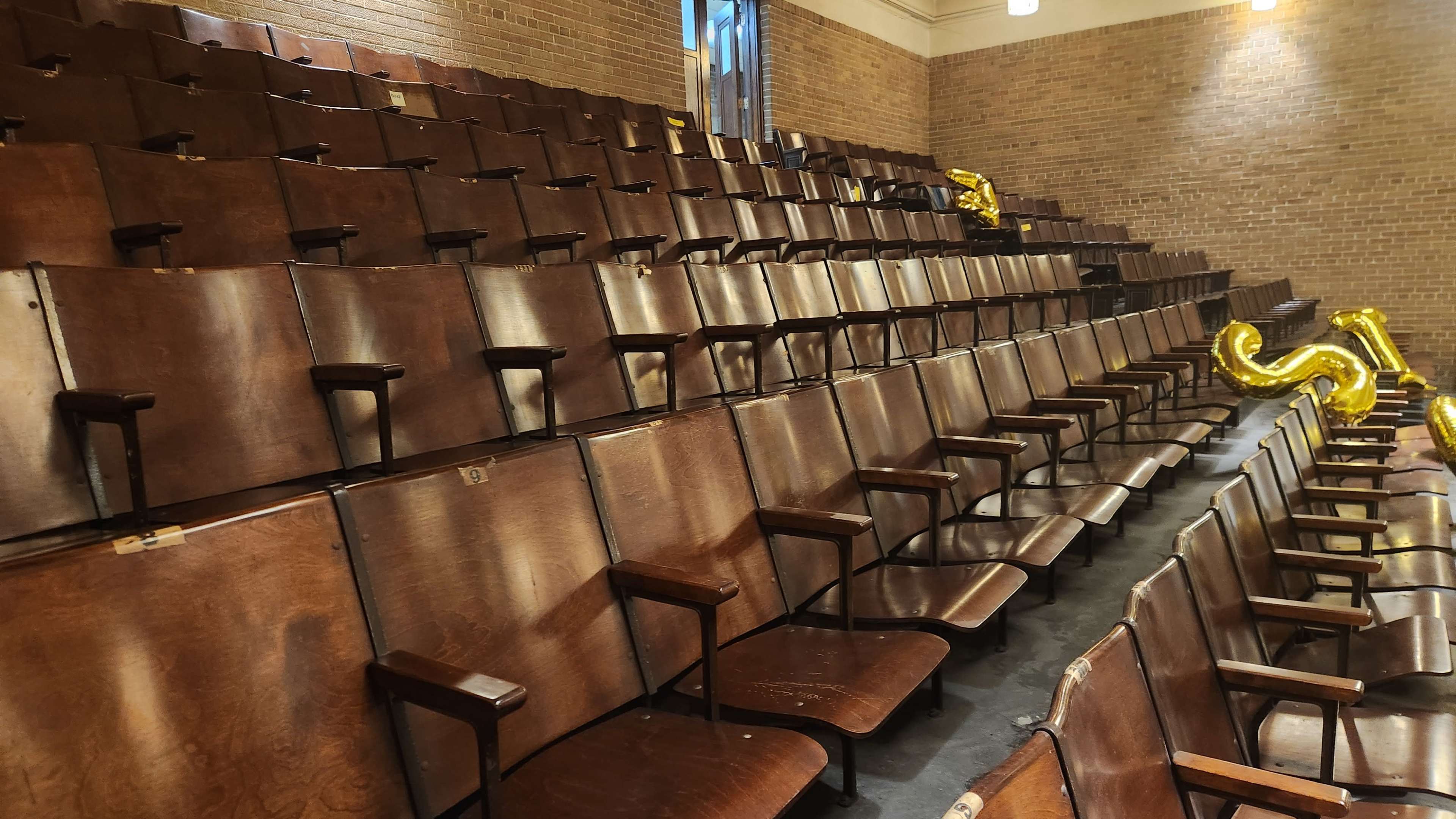 The image shows an empty auditorium with wooden seats arranged in rows, highlighting a brick wall in the background.
