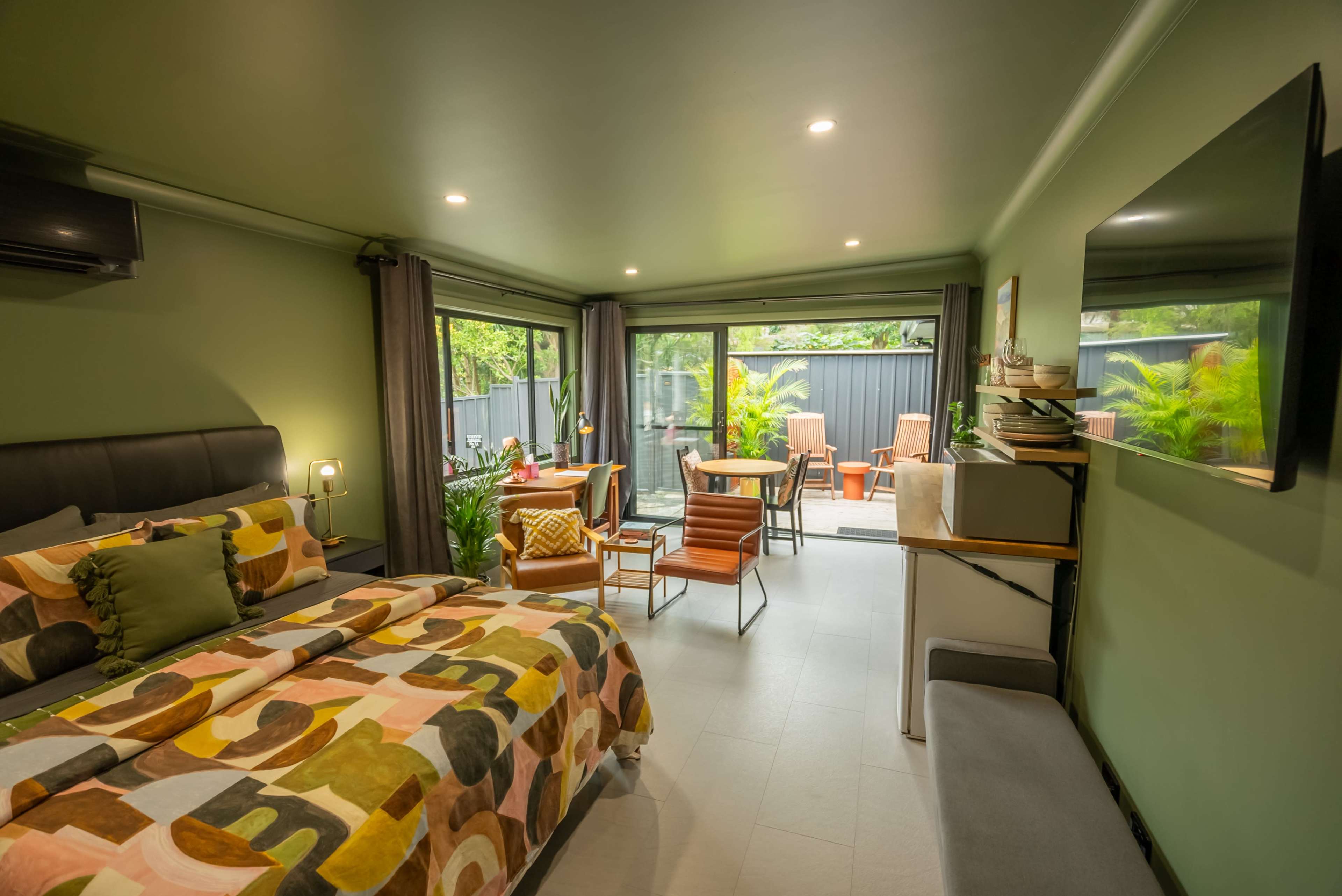 A modern bedroom features a bed with a colorful patterned duvet, seating areas, and large windows that open to a patio with outdoor furniture.