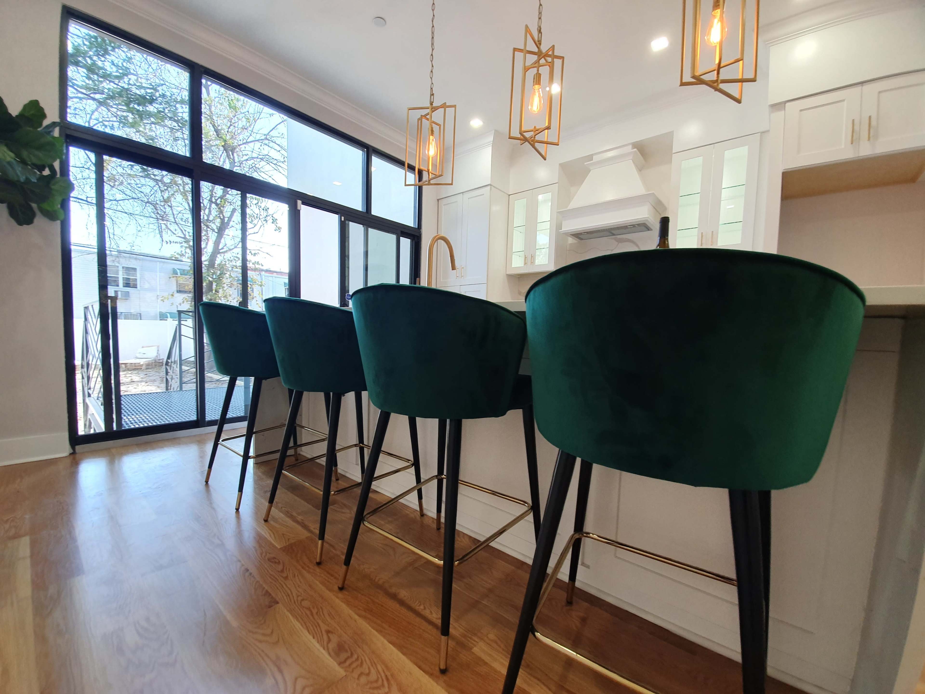 A modern kitchen with four green velvet bar stools lined up against a countertop, illuminated by pendant lights.