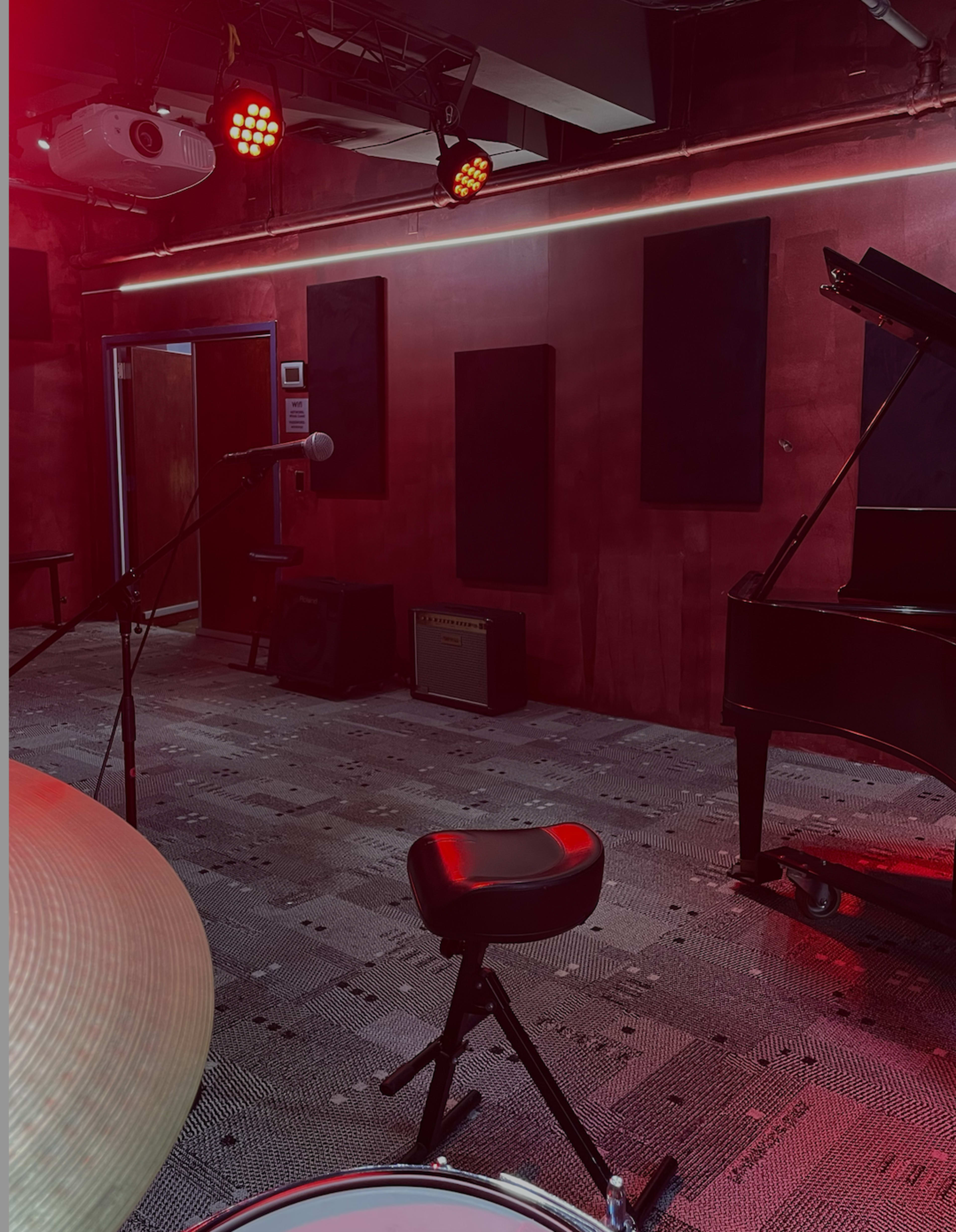 The image shows a music rehearsal room featuring a grand piano, a microphone on a stand, and stage lights mounted on the ceiling.