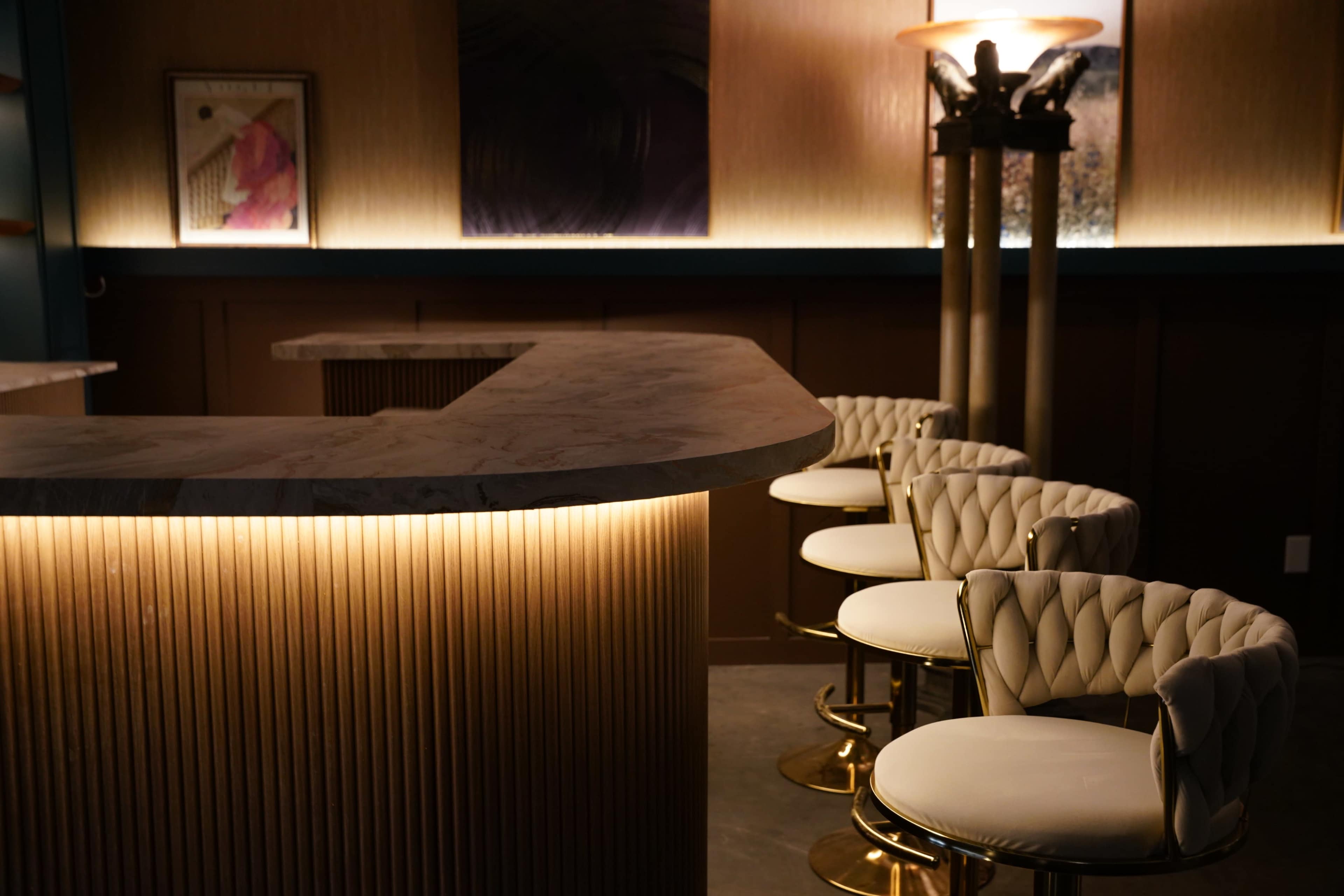 The image shows a sleek, modern bar with a marble countertop and upholstered stools illuminated by soft lighting.