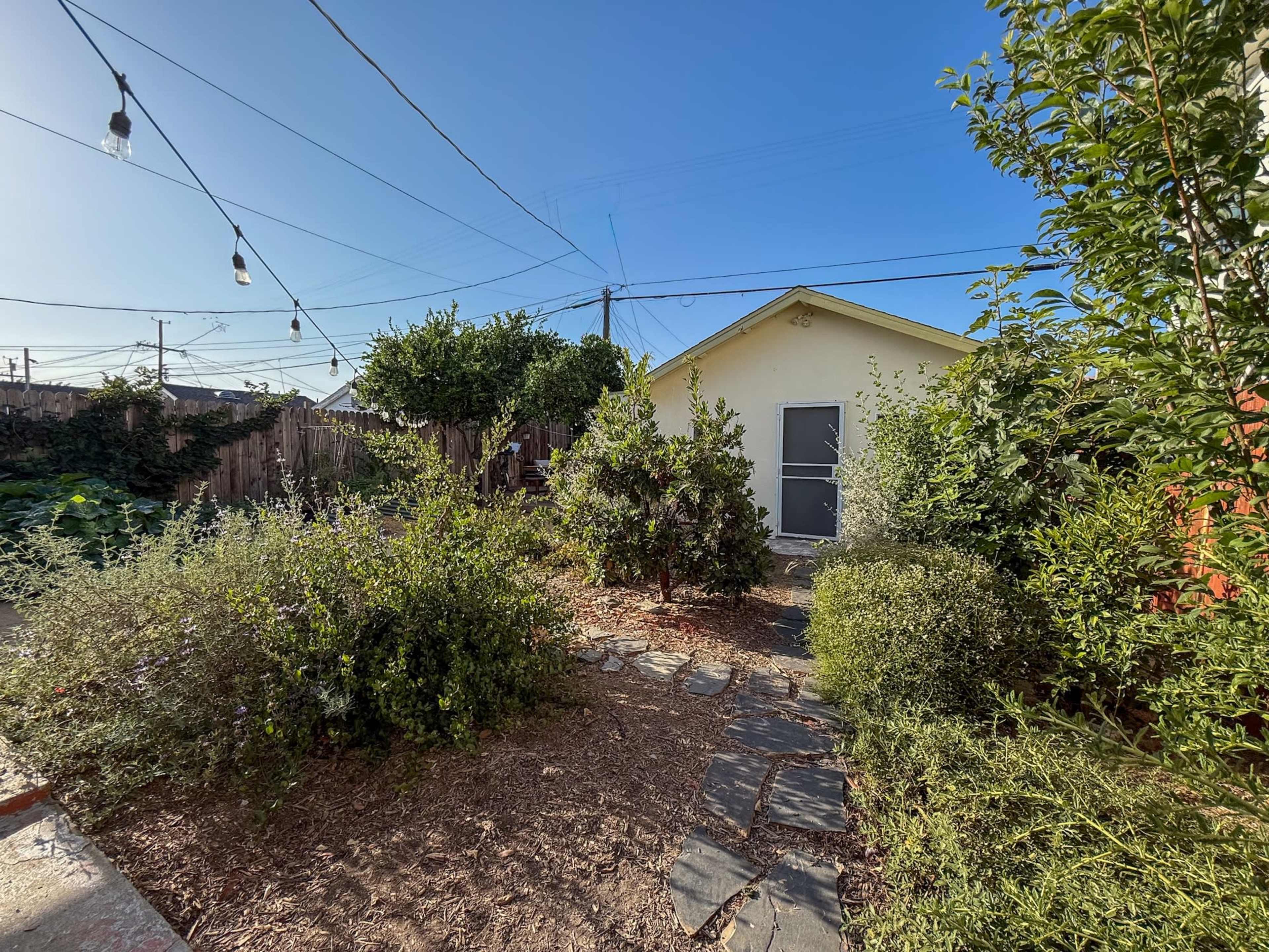 The image shows a backyard scene with a path made of stones leading to a yellow shed, surrounded by various plants and trees under a clear blue sky.