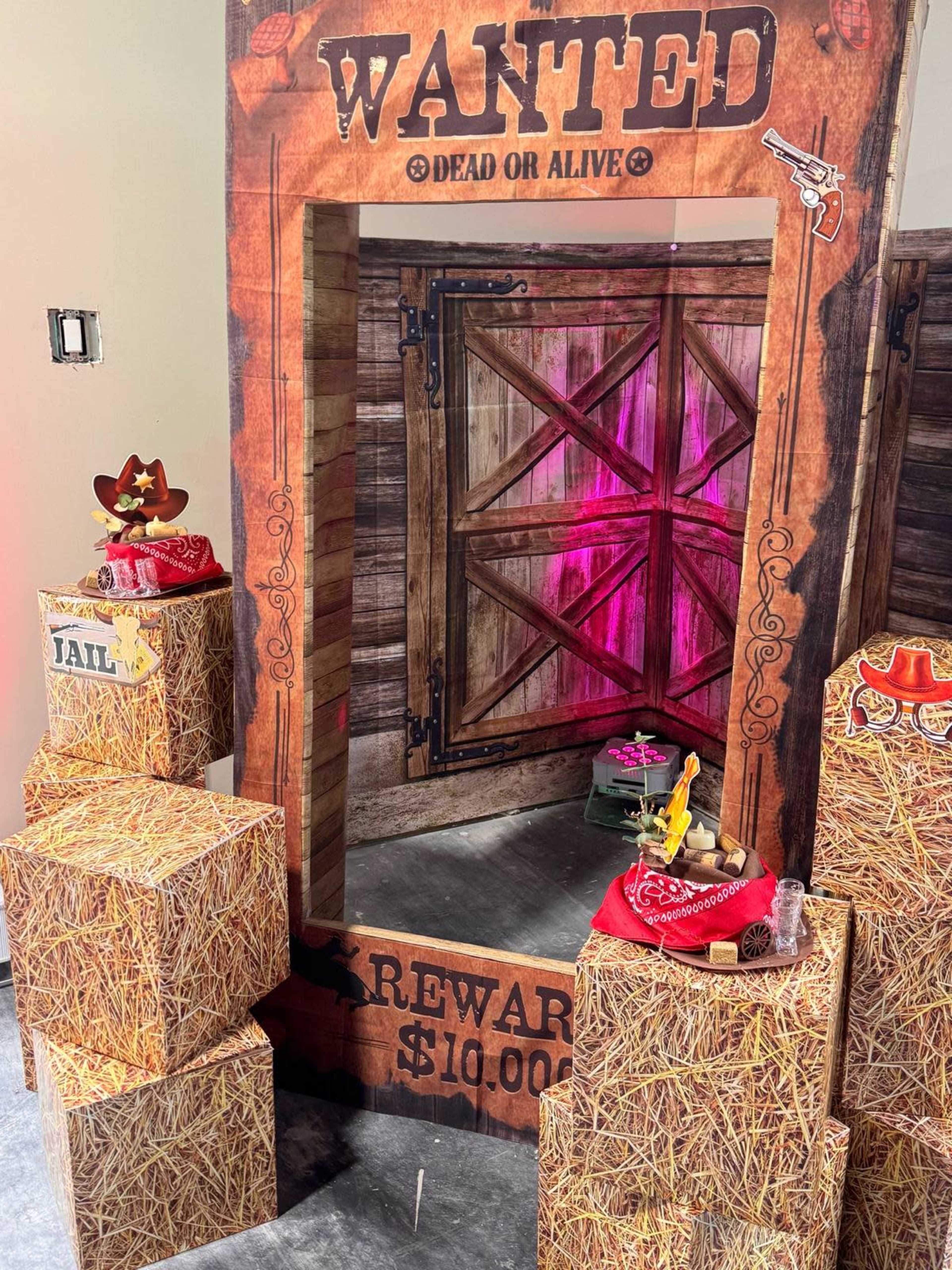 The image shows a western-themed photo booth featuring a large "WANTED" sign, a wooden door backdrop, and hay bale decorations.