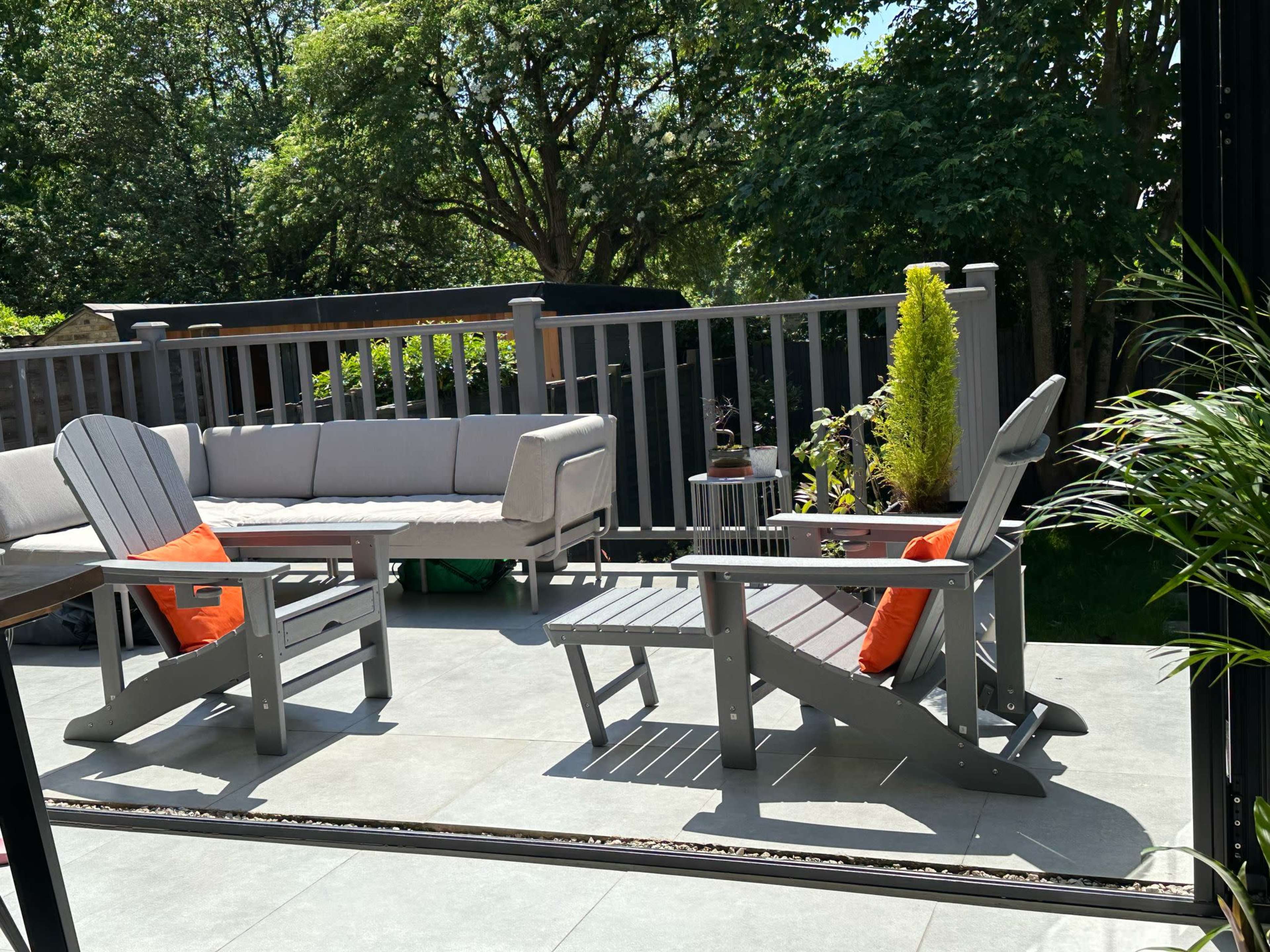 A patio features two gray Adirondack chairs with orange cushions, a light gray sofa, and a wooden railing surrounded by lush greenery.