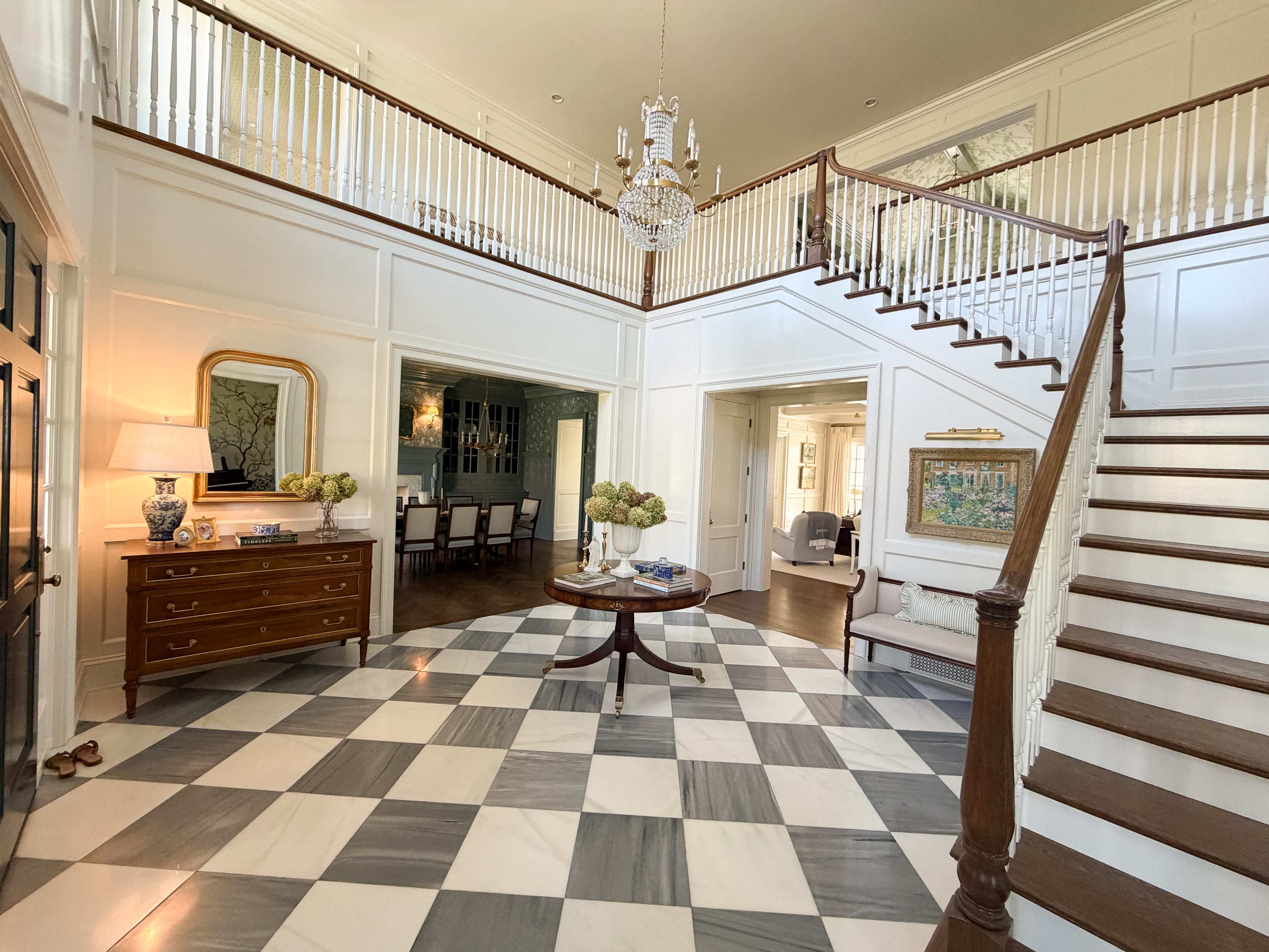 The entrance hall features a checkered marble floor, a staircase with wooden railings, and a crystal chandelier.