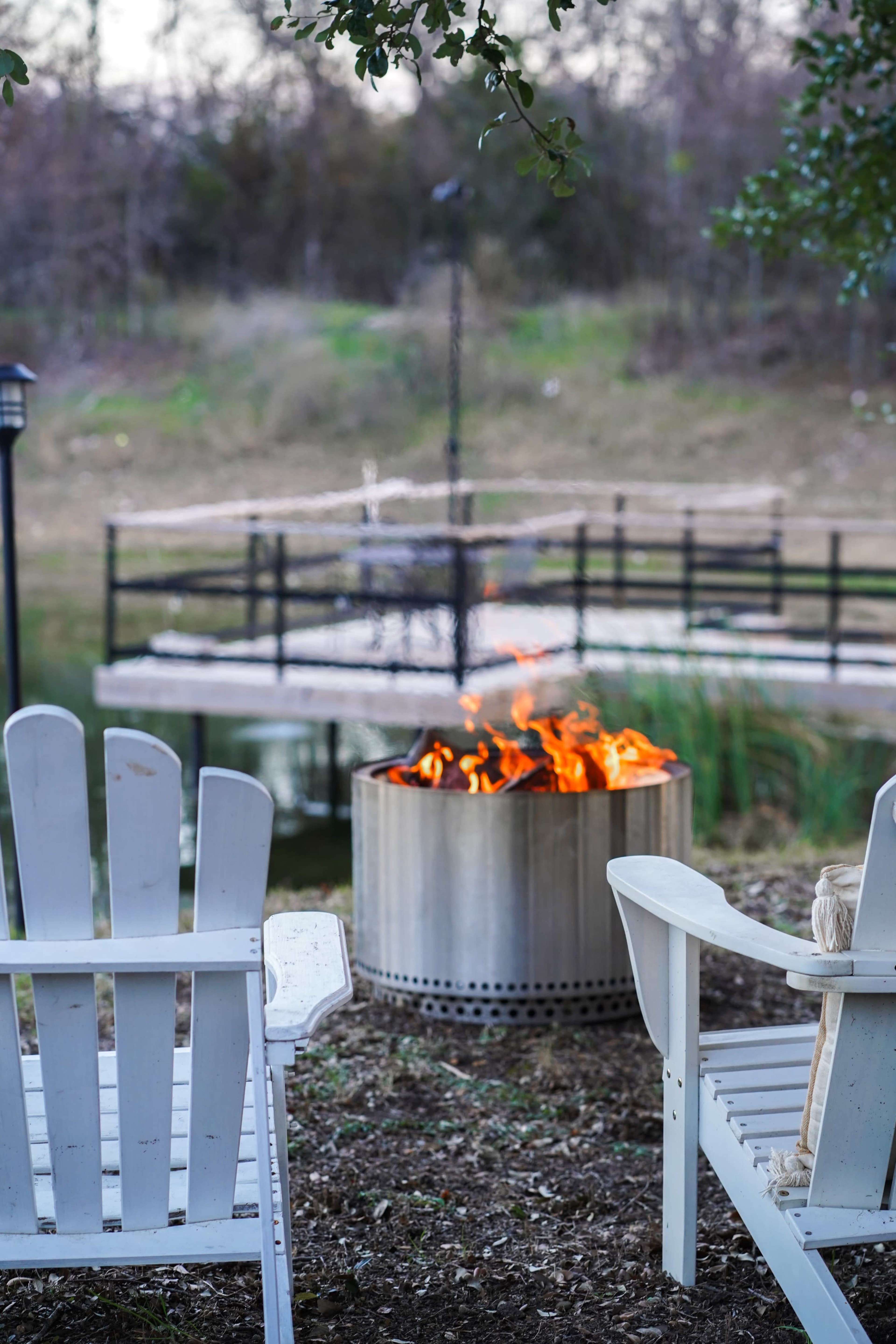 A fire burns in a metal fire pit surrounded by two white Adirondack chairs near a pond with a small dock in the background.
