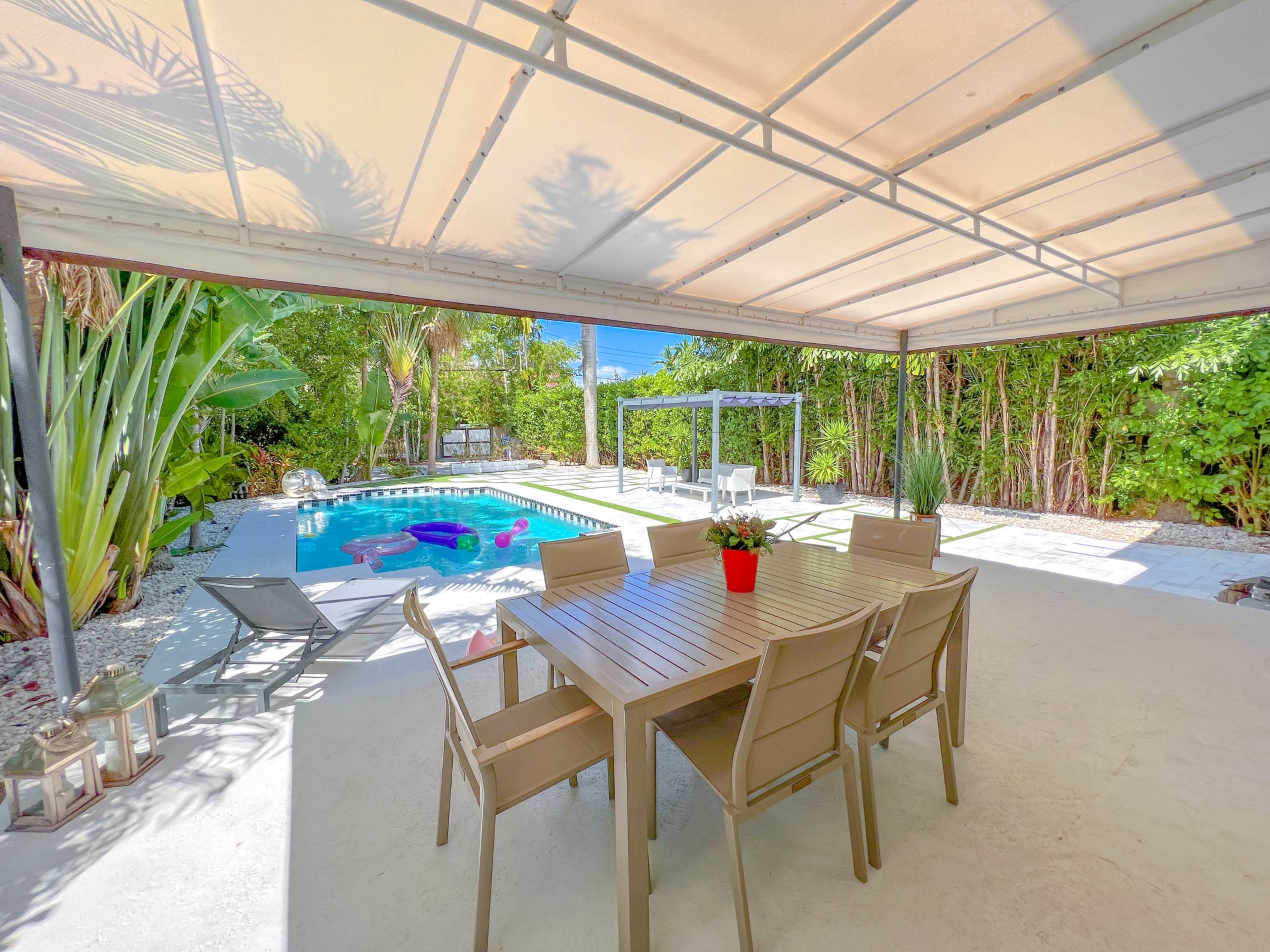 A patio with a dining table and chairs overlooks a pool surrounded by tropical plants.