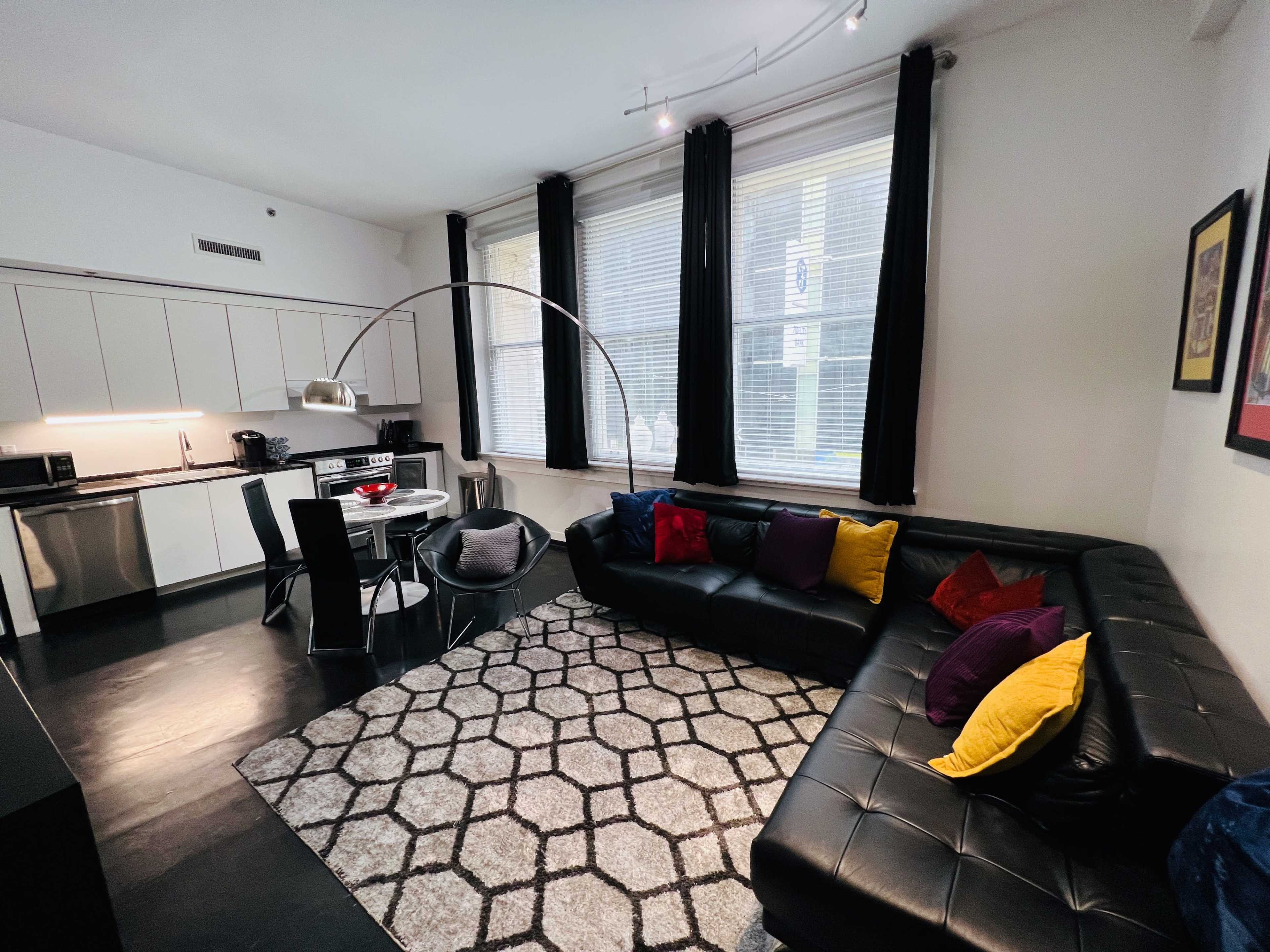 The image shows a modern apartment living space with a black leather sectional, a round dining table, and large windows, adorned with a geometric rug and colorful pillows.