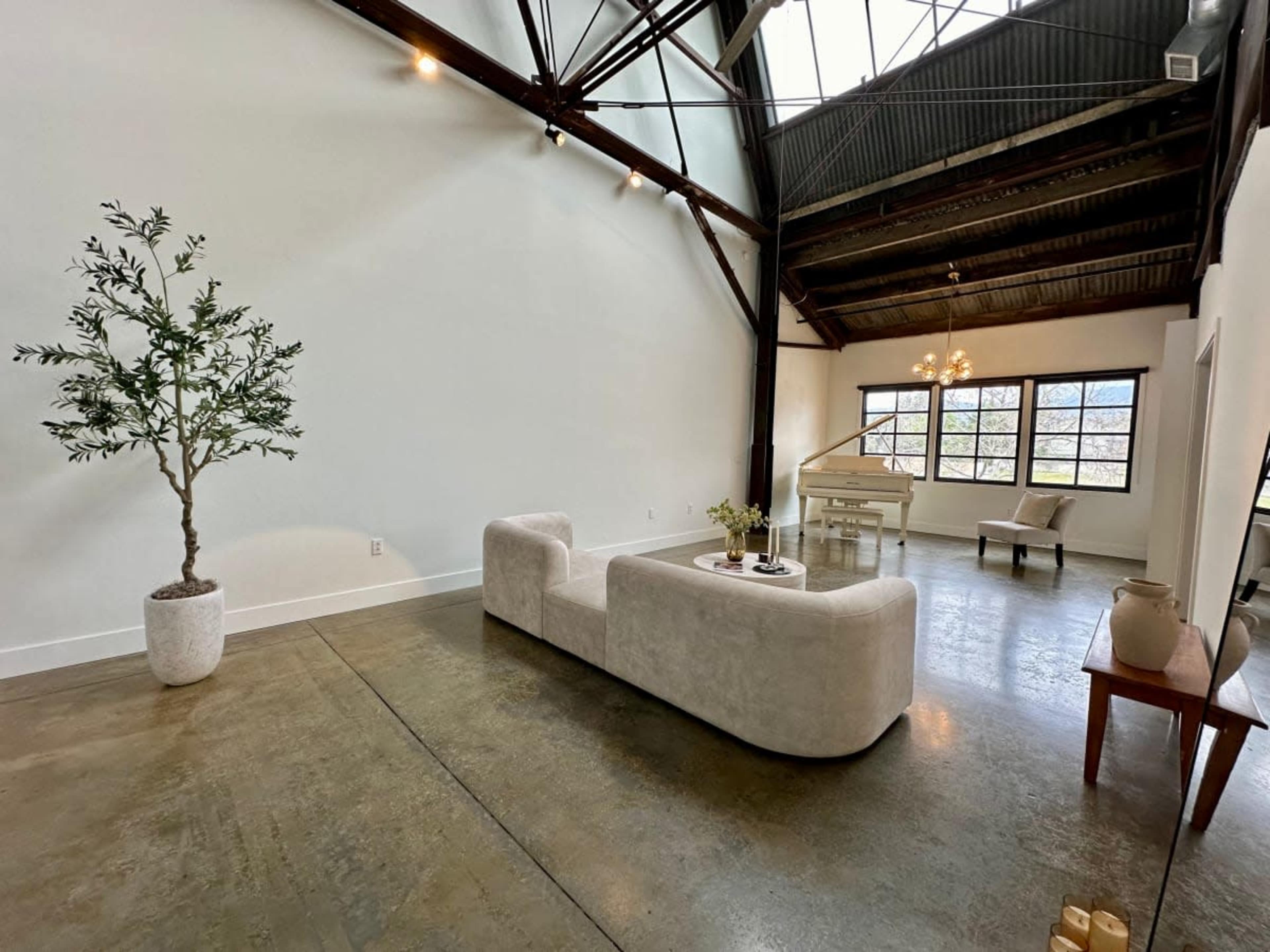 The image shows a spacious, industrial-style interior with a large sectional sofa, a dining area, and a potted plant, all under a high ceiling with exposed beams and large windows.