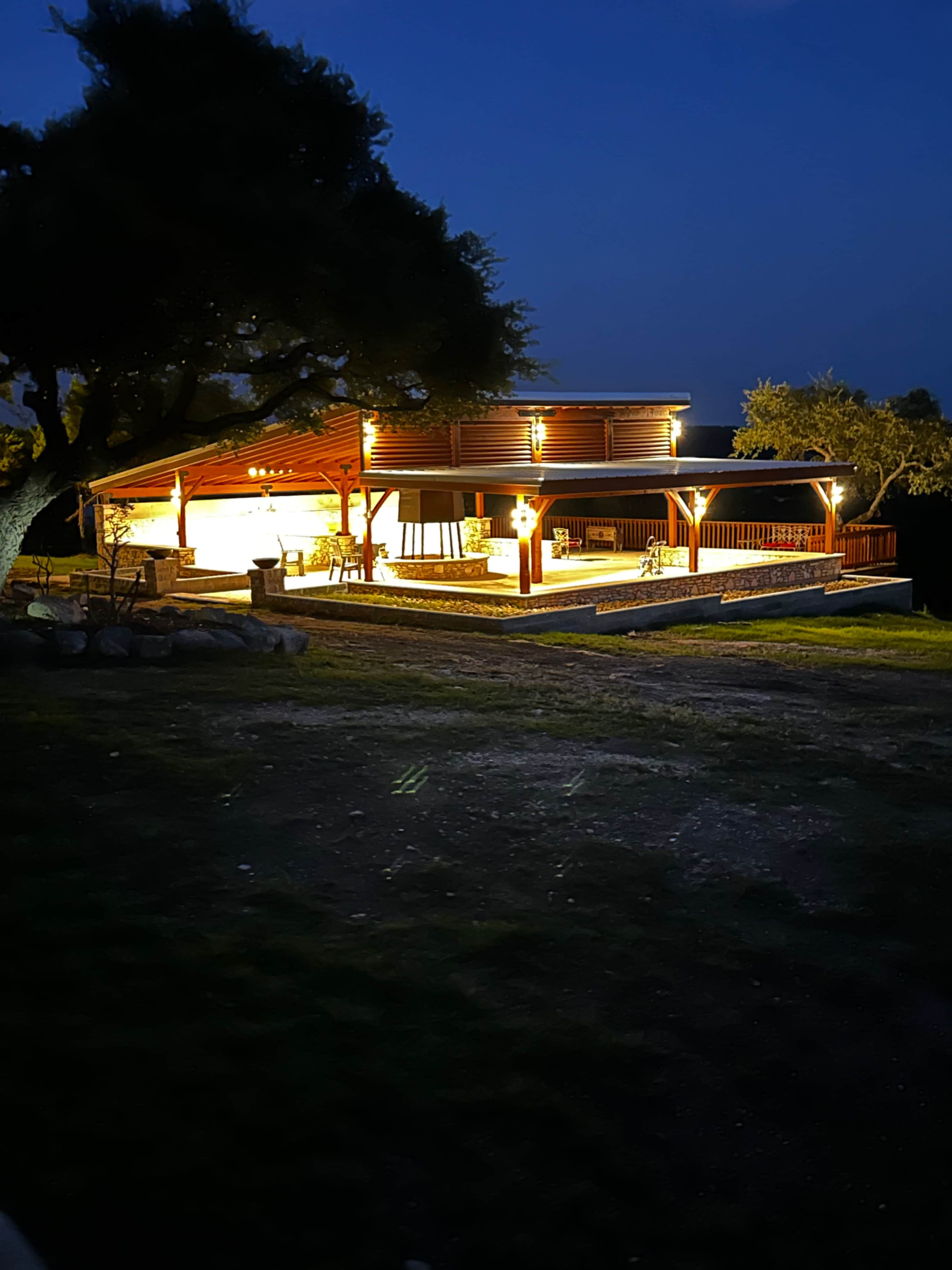 A wooden pavilion with warm lights stands in a grassy area at night, surrounded by trees.
