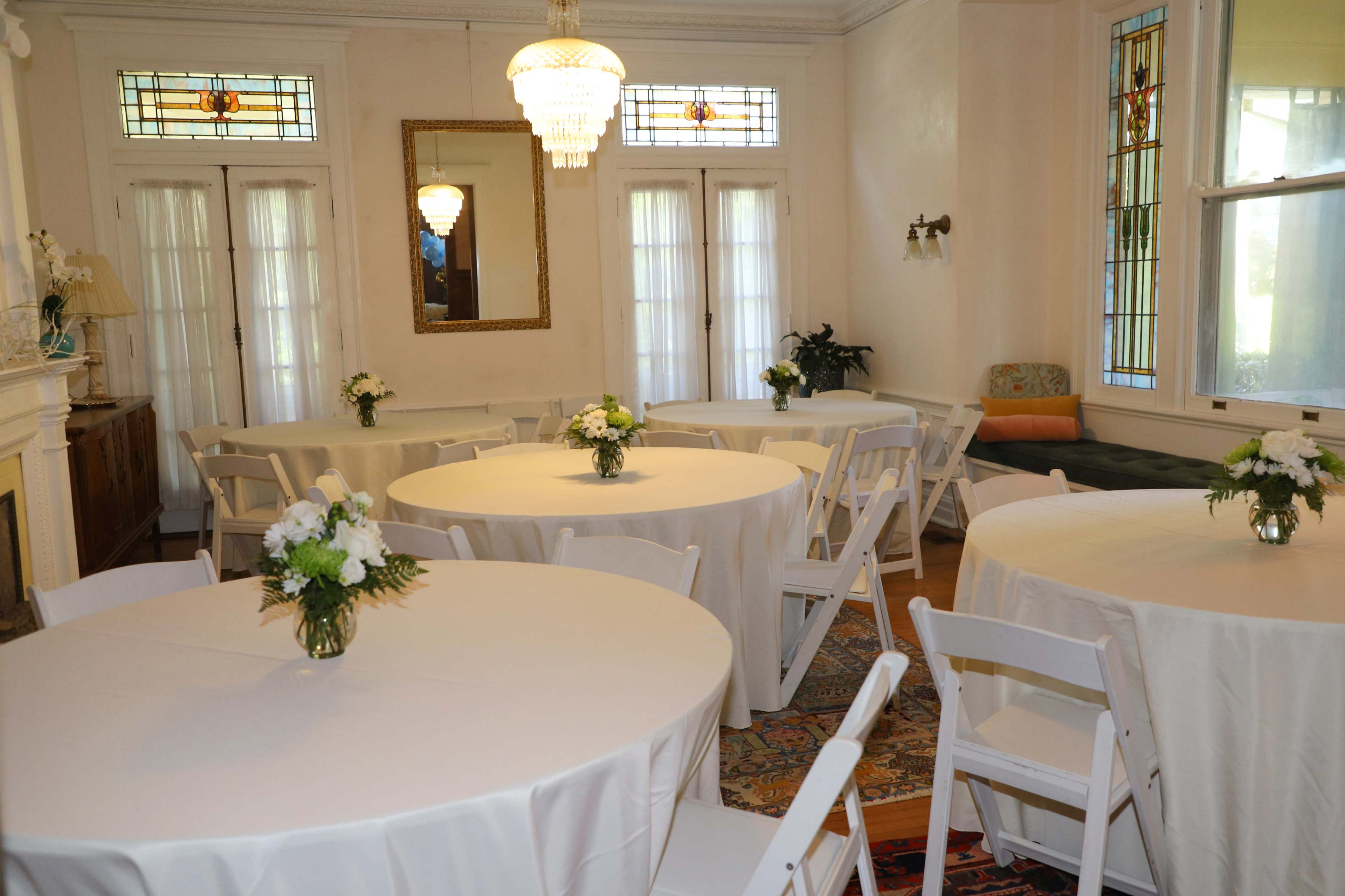 A room with four round tables covered in white tablecloths, each adorned with small floral centerpieces, in a well-lit space featuring large windows and a chandelier.