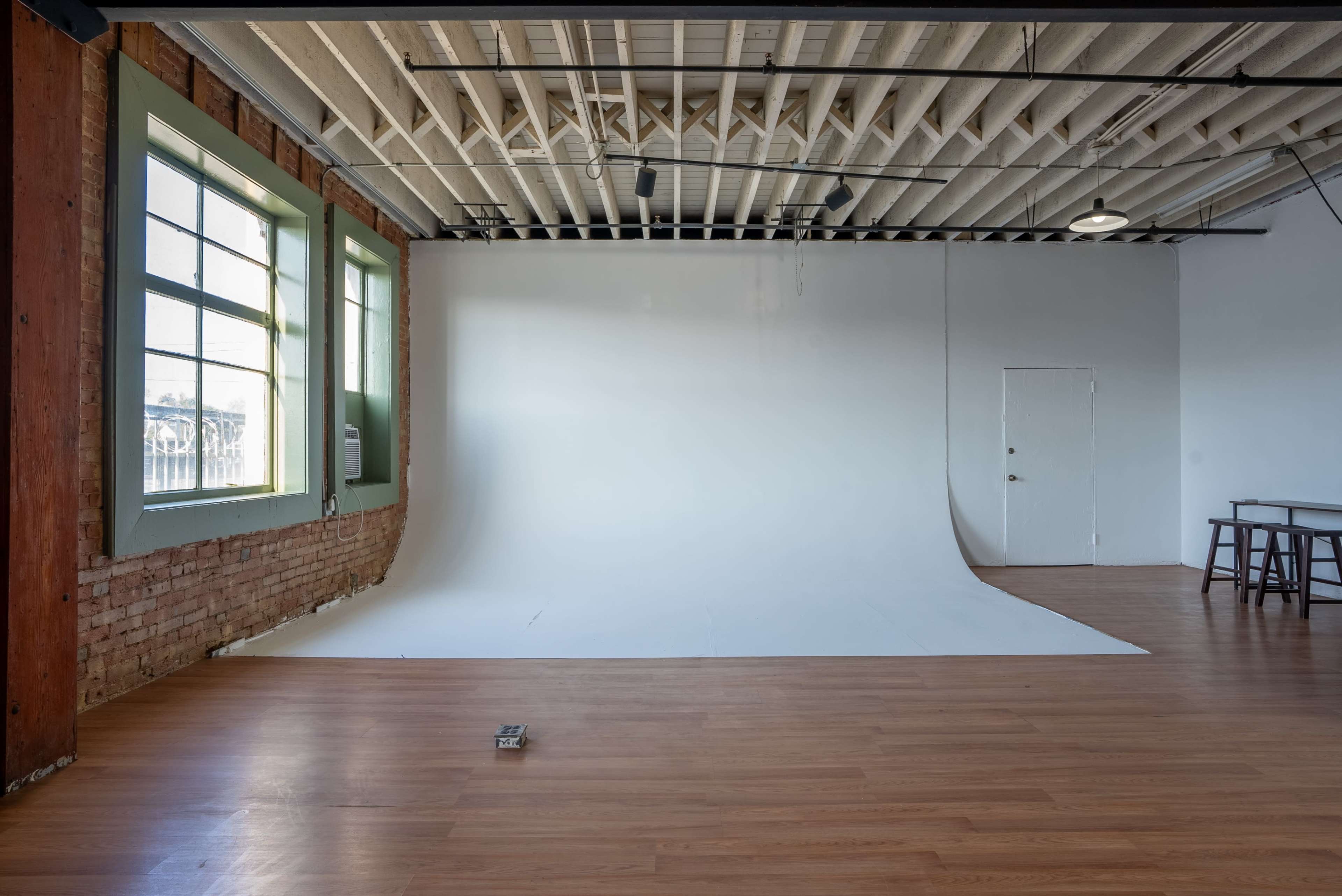 The image shows a spacious photography studio with a white backdrop, large windows, wooden flooring, and exposed brick walls.