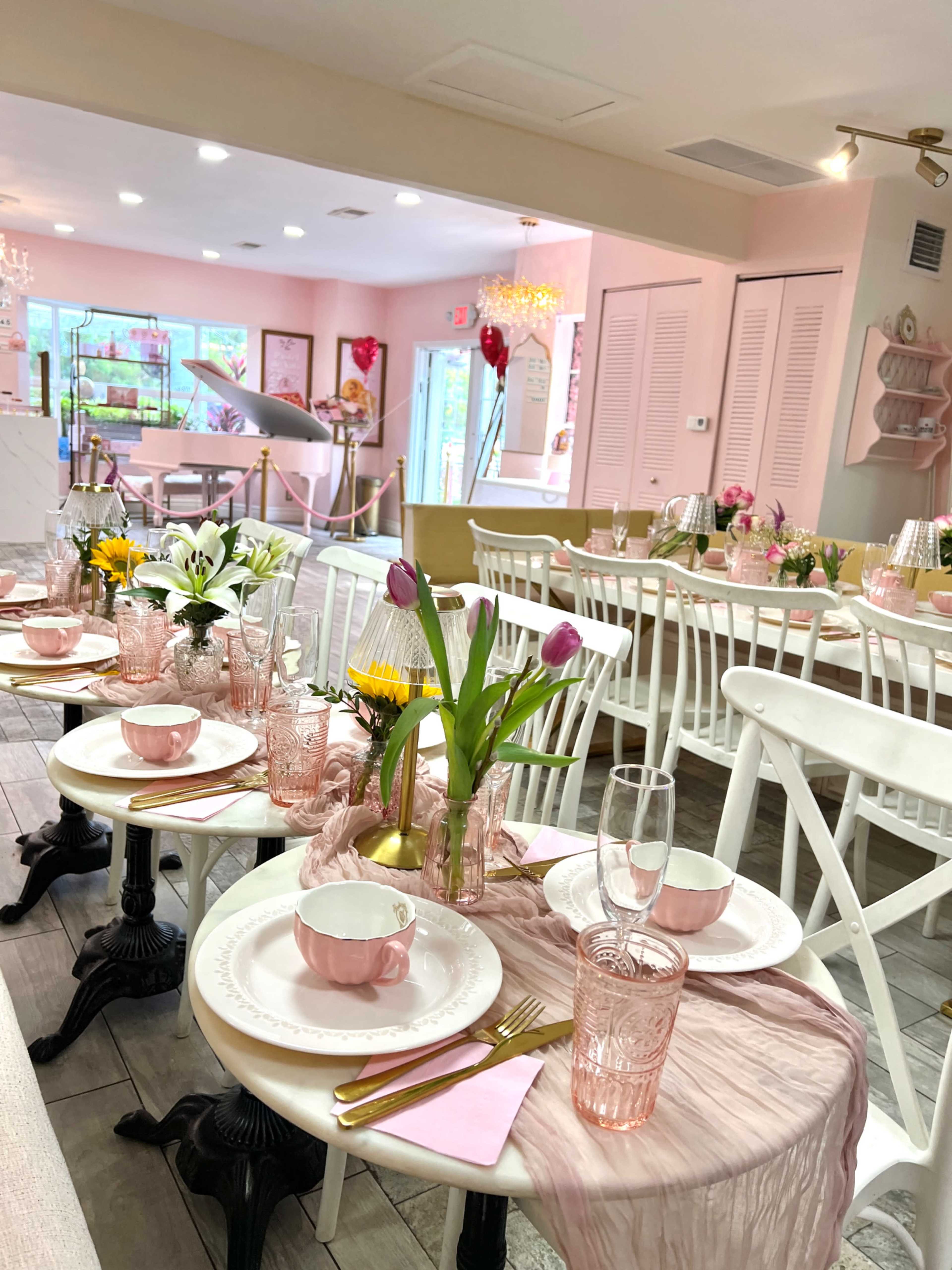 A bright, elegantly decorated dining area features tables set with pink dishes, glassware, and fresh flowers, creating a welcoming atmosphere.